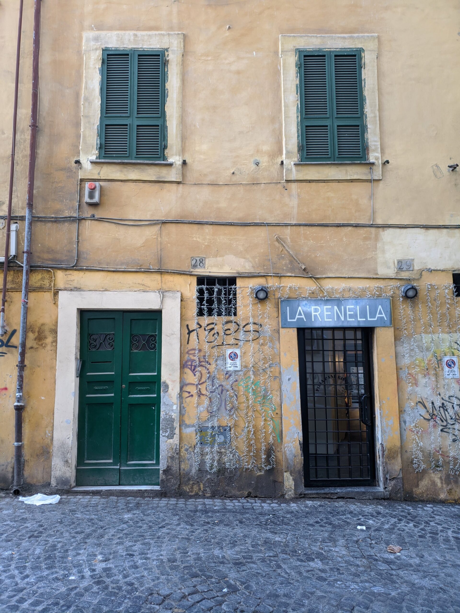 Fachada de um edifício em Trastevere com portas verdes, janelas com persianas fechadas e paredes desgastadas pela idade, exibindo um charme urbano e histórico, com grafites e plantas que decoram o exterior.