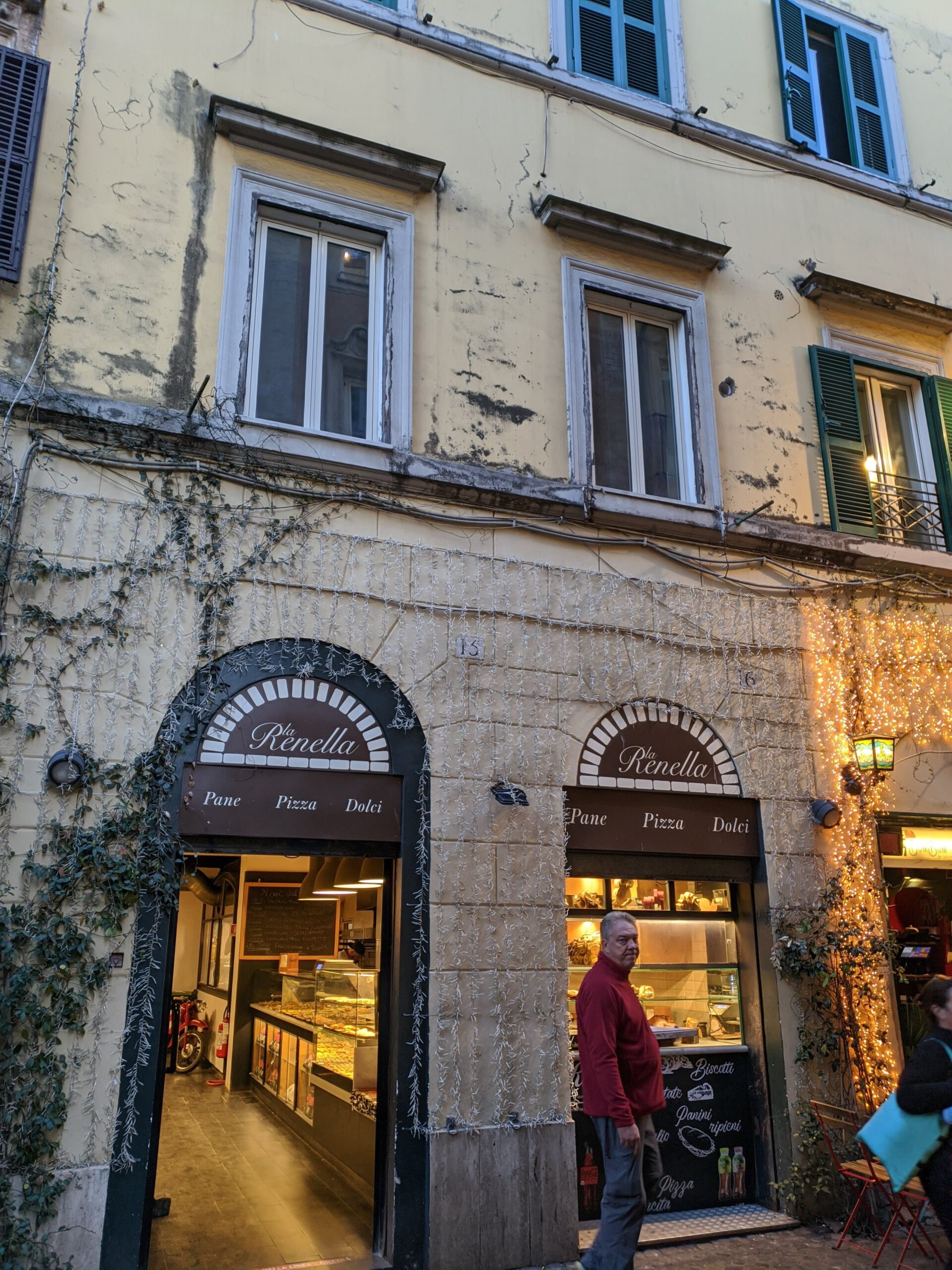 Fachada da famosa padaria Renella em Trastevere, com sua entrada arqueada e decoração luminosa, exibindo uma atmosfera acolhedora e rústica, enquanto um homem caminha por sua frente.