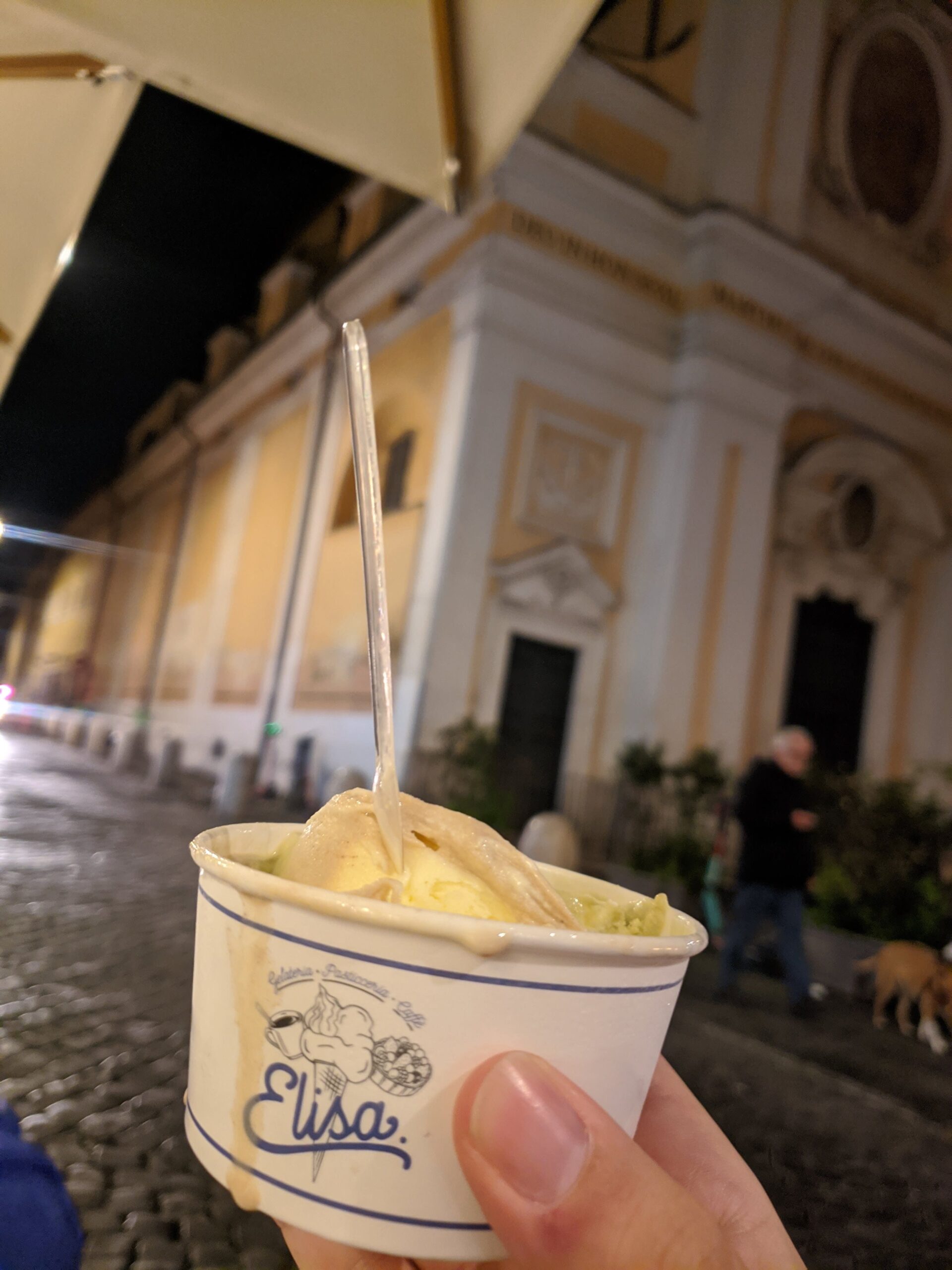 Uma mão segurando um copo de papel com gelato de pistache e baunilha, com uma colher branca espetada, contra o fundo desfocado de uma praça noturna em Roma.