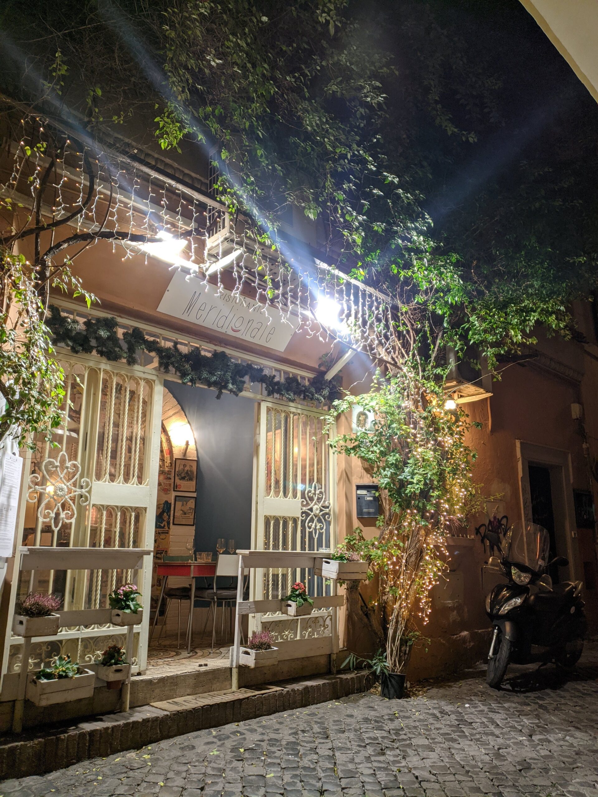 Uma pequena entrada de restaurante em Trastevere, iluminada por luzes decorativas que destacam a vegetação e a arquitetura tradicional, com uma motocicleta estacionada ao lado, criando um ambiente convidativo e intimista.