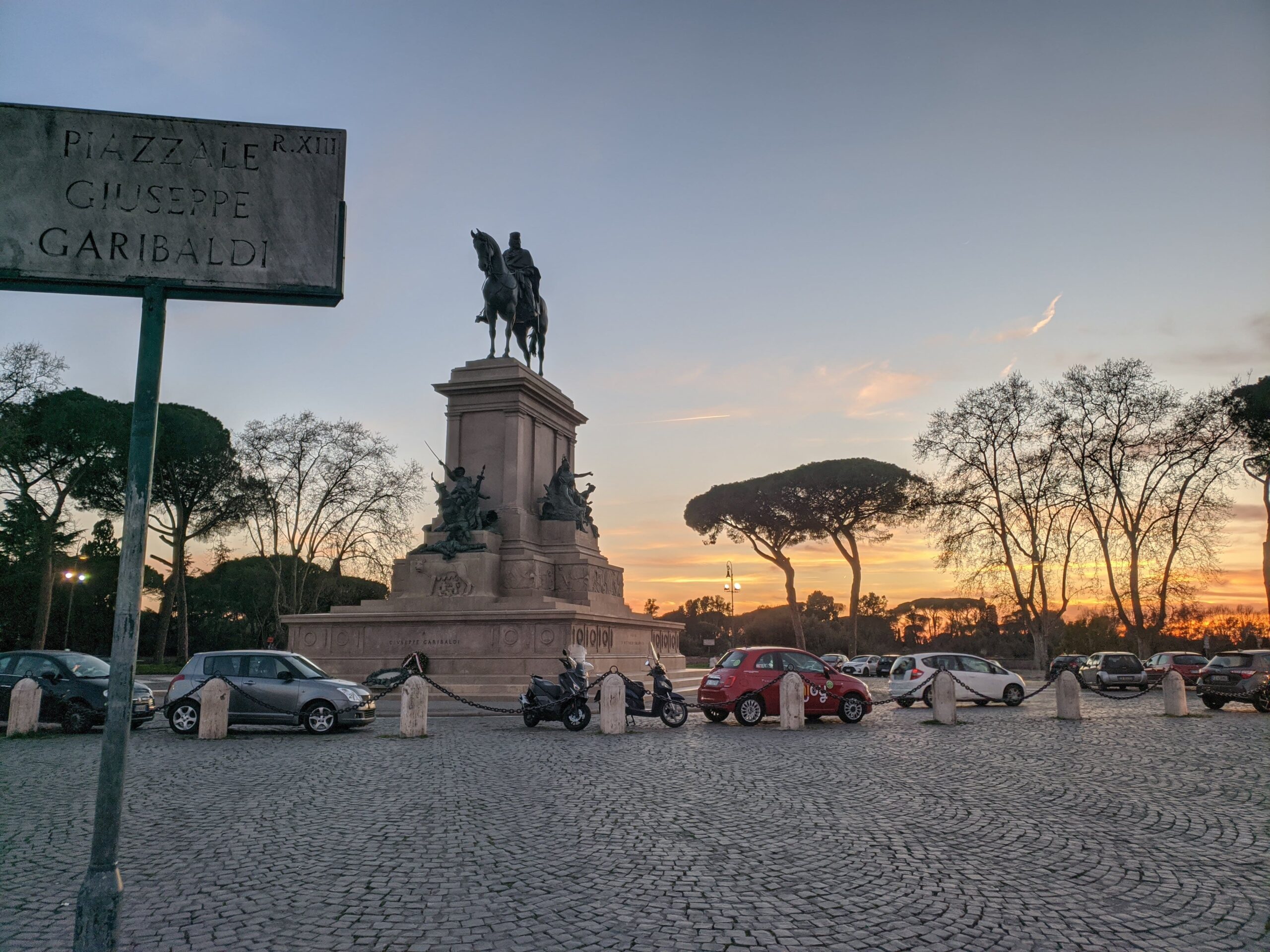Monumento ao entardecer, com a estátua de Giuseppe Garibaldi montado em um cavalo se destacando contra um céu colorido pelo pôr do sol, localizado em uma praça pavimentada com carros estacionados e pinheiros-marítimos ao fundo.