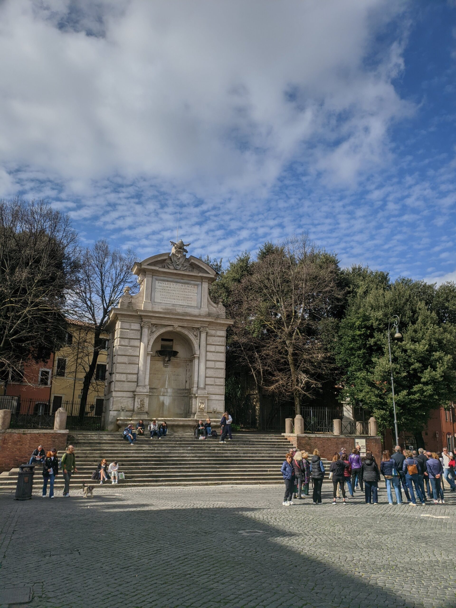 Piazza Trilussa em Roma, dia claro com céu parcialmente nublado, uma fonte histórica ao centro, rodeada por árvores e pessoas sentadas em seus degraus, desfrutando de um momento de descanso.