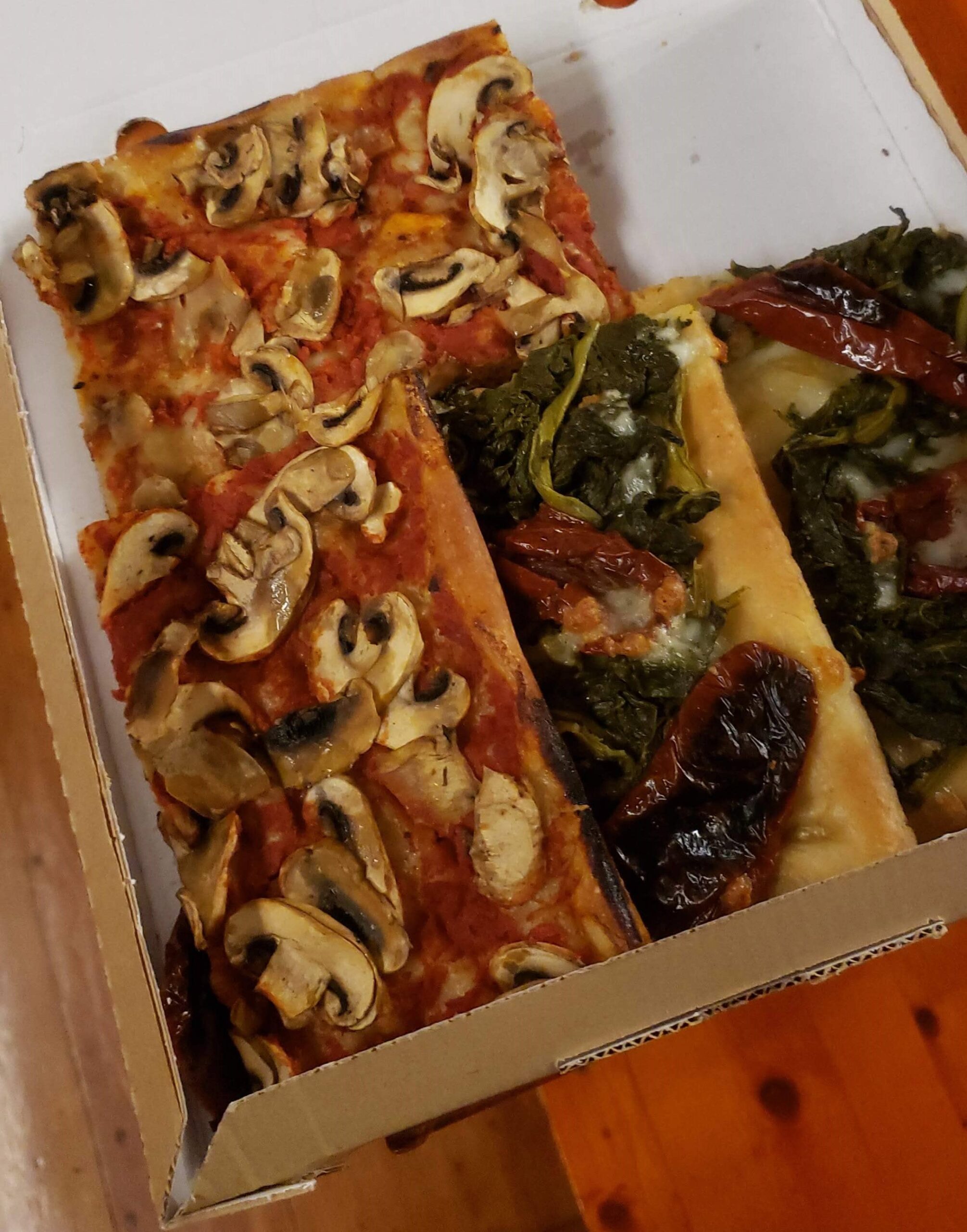 Uma fatia de pizza com cogumelos e outra com espinafre e tomate seco em uma caixa aberta sobre uma mesa de madeira.