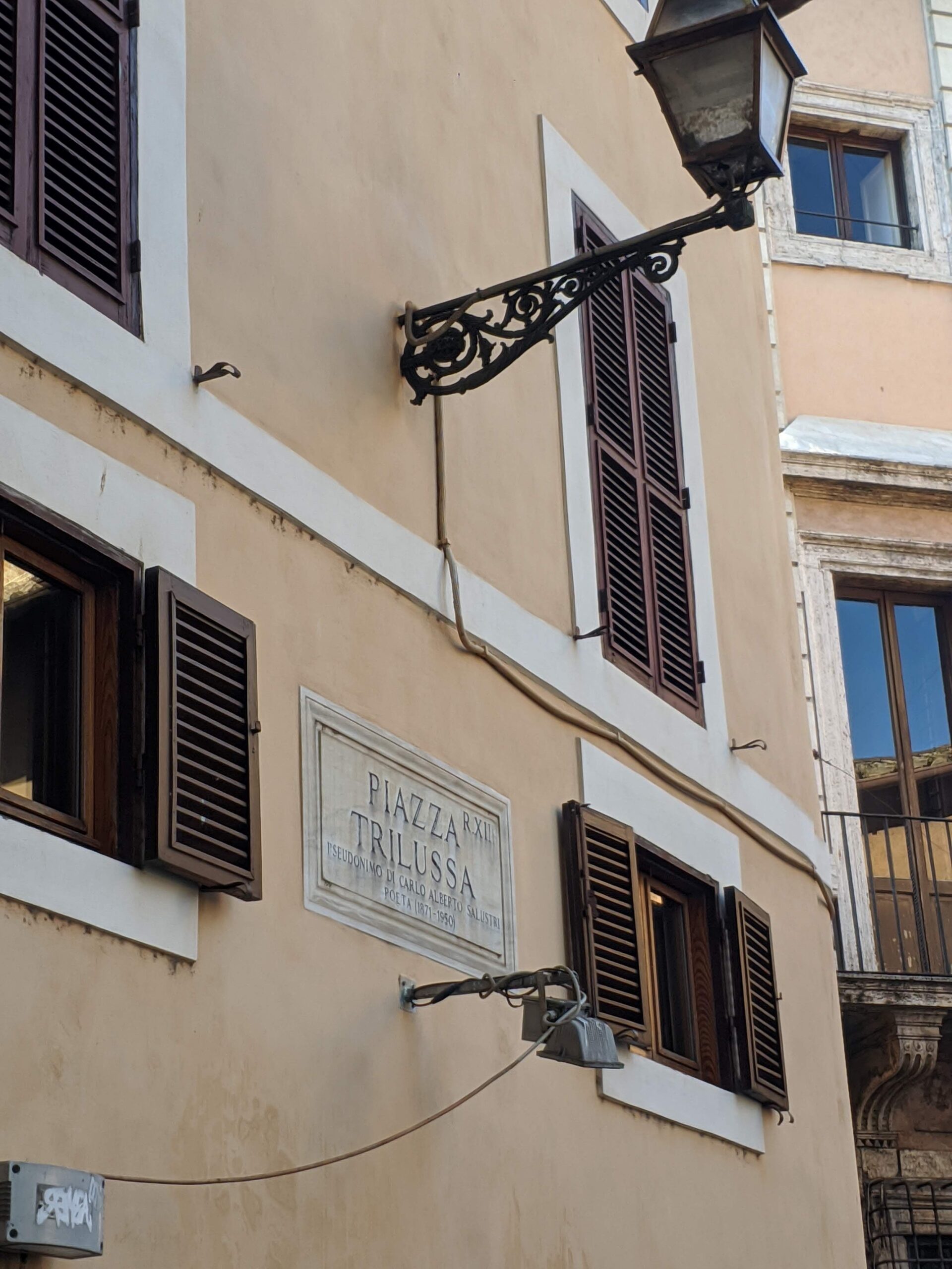 Placa de rua branca com a inscrição "PIAZZA TRILUSSA" em letras pretas, fixada na parede de um edifício antigo com janelas de persianas fechadas, com um poste de luz de metal ornamentado à frente.