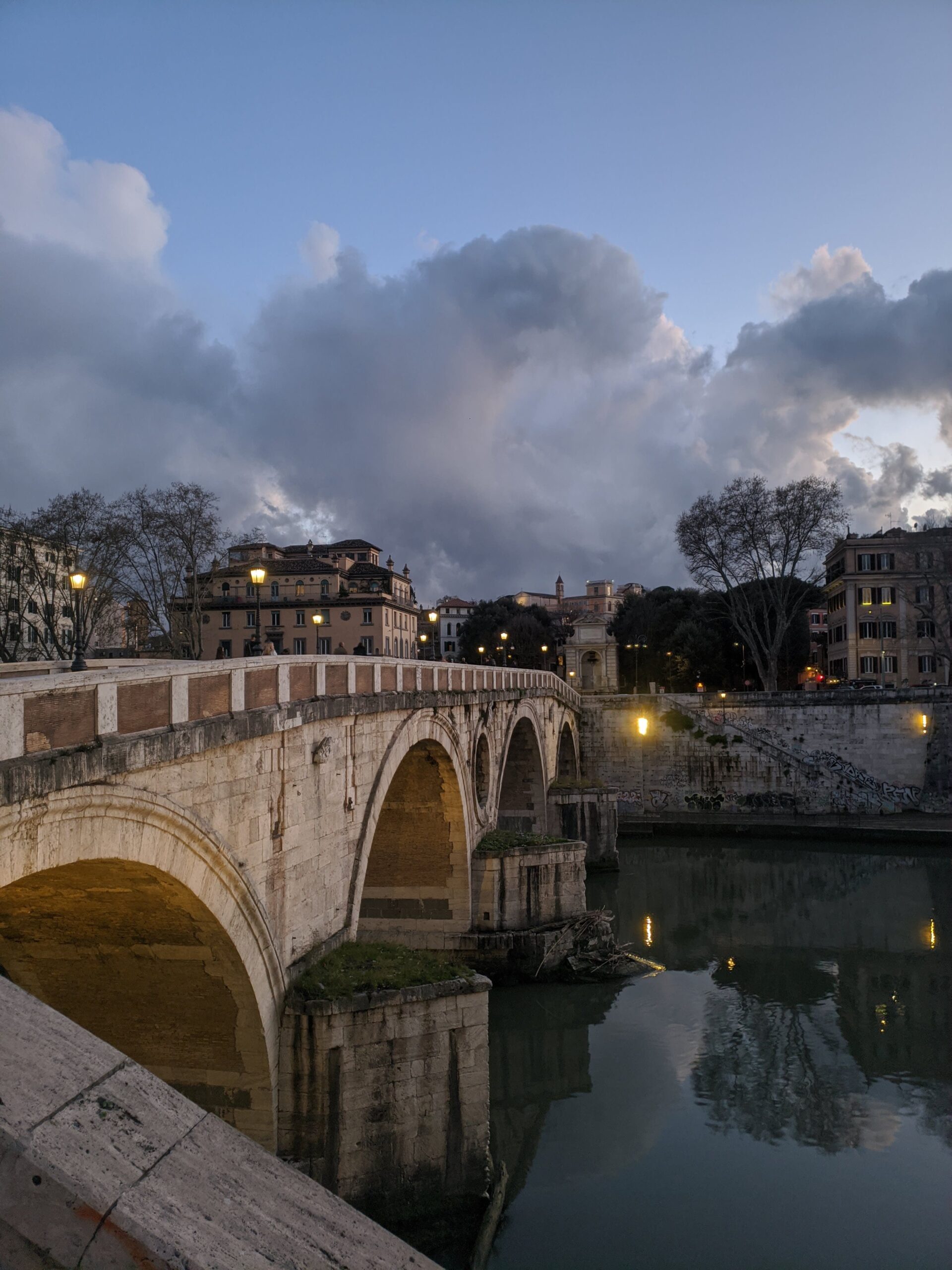 A histórica Ponte Sisto em Roma durante o crepúsculo, com o céu dramaticamente nublado refletido sobre as águas tranquilas do rio Tibre, enquanto as luzes da cidade começam a se acender, destacando a beleza da arquitetura e do ambiente ao entardecer.