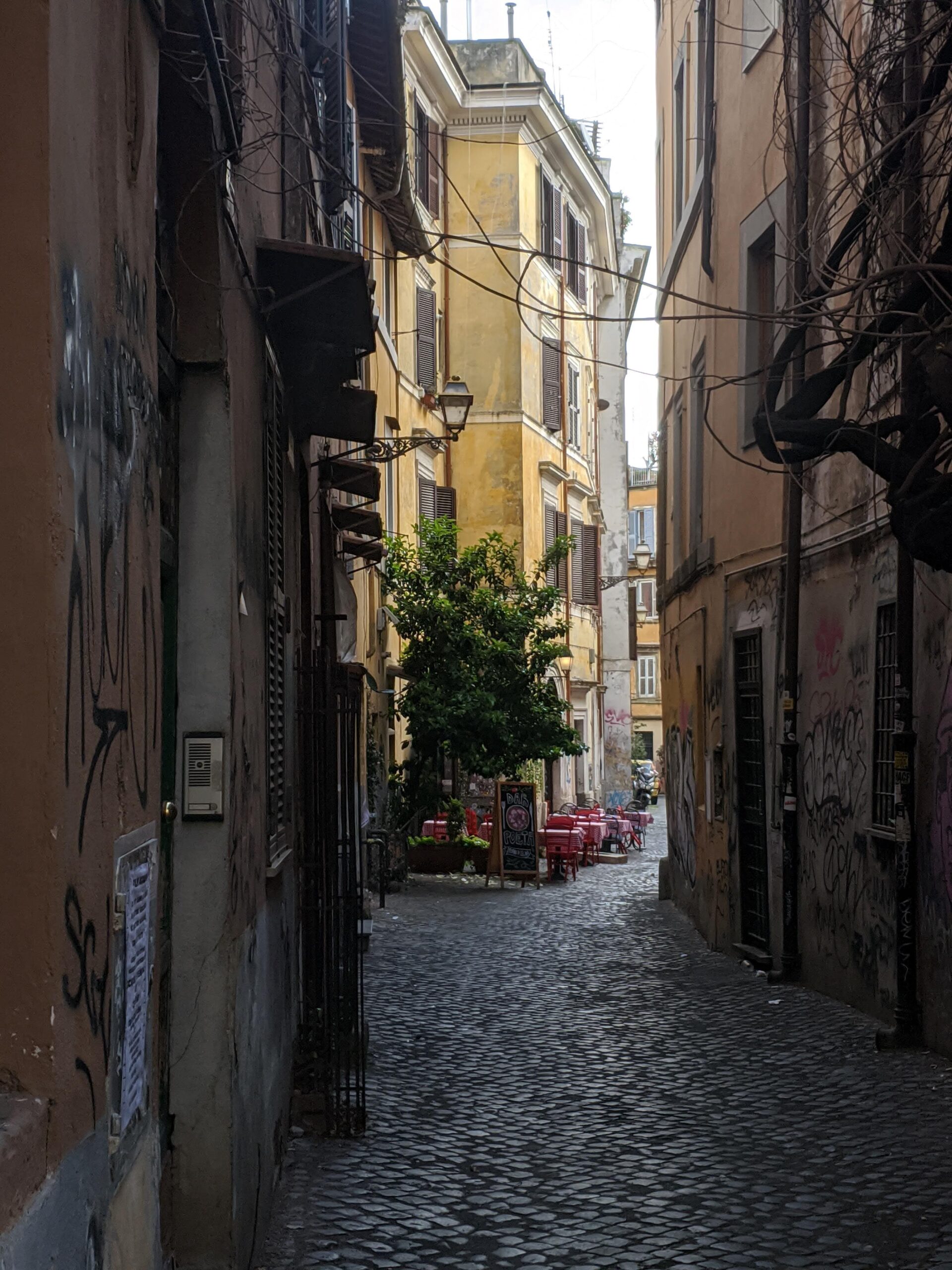 Rua de paralelepípedos em Trastevere ao amanhecer, com edifícios históricos desgastados pelas intempéries e a tranquilidade de uma manhã sem a agitação costumeira, apenas algumas mesas de café dispostas na calçada.