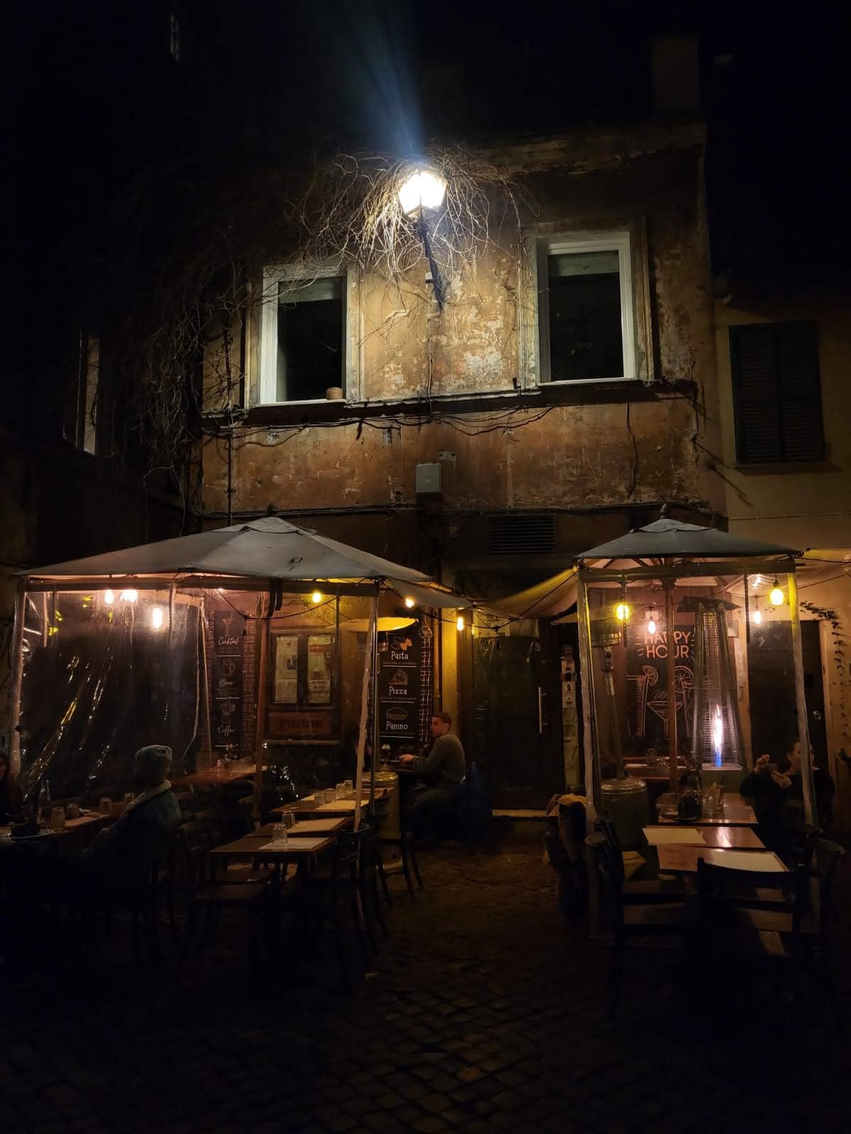 Cena noturna de um restaurante ao ar livre com mesas vazias e uma iluminação suave, em frente a uma fachada rústica e envelhecida, transmitindo uma atmosfera acolhedora e tranquila no bairro Trastevere em Roma.