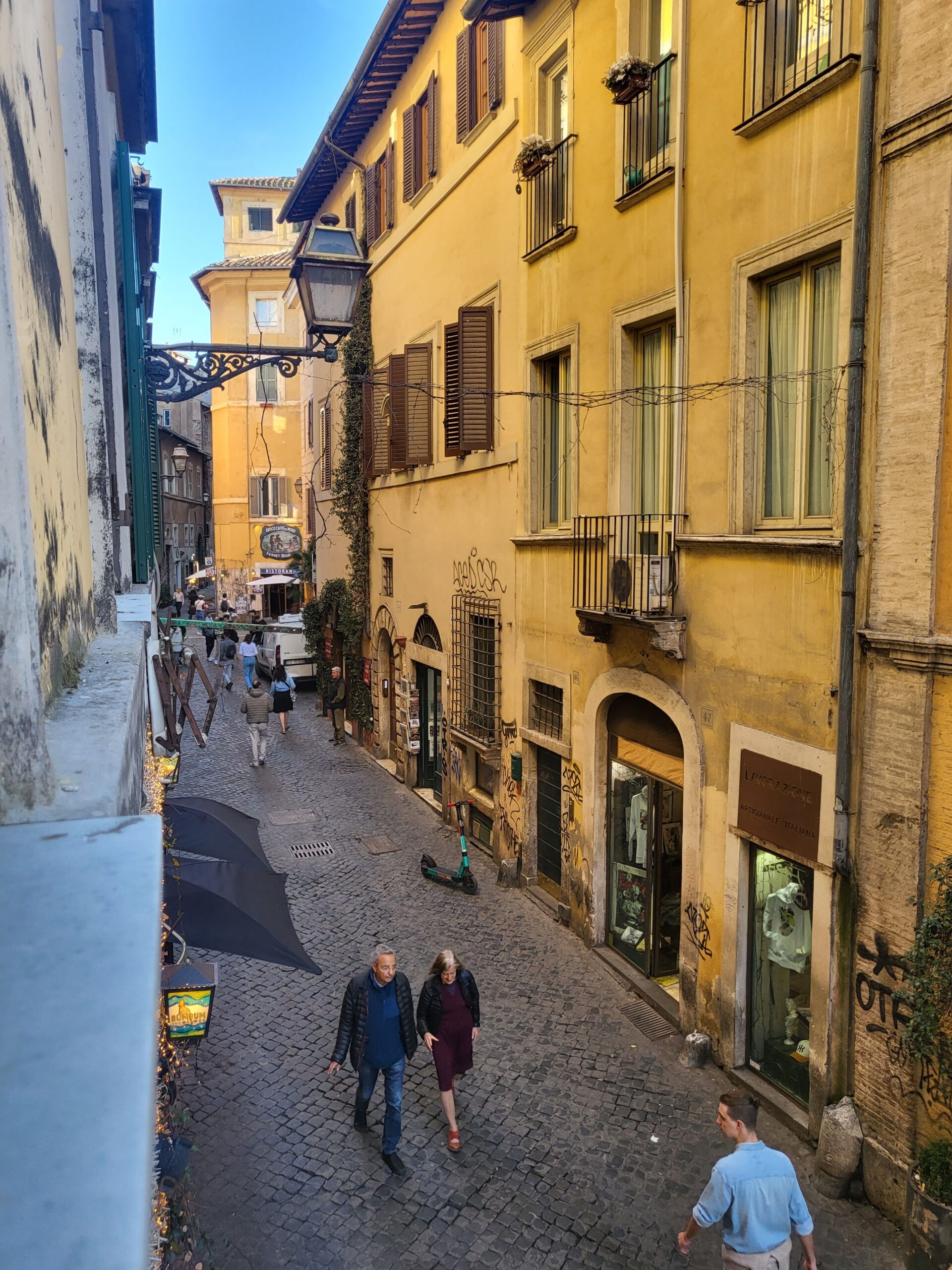 Vista diurna de um ponto elevado em Trastevere, Roma, exibindo a arquitetura histórica com edifícios em tons de ocre e janelas com venezianas, uma rua estreita abaixo movimentada por pedestres, oferecendo um vislumbre da vida cotidiana na cidade.