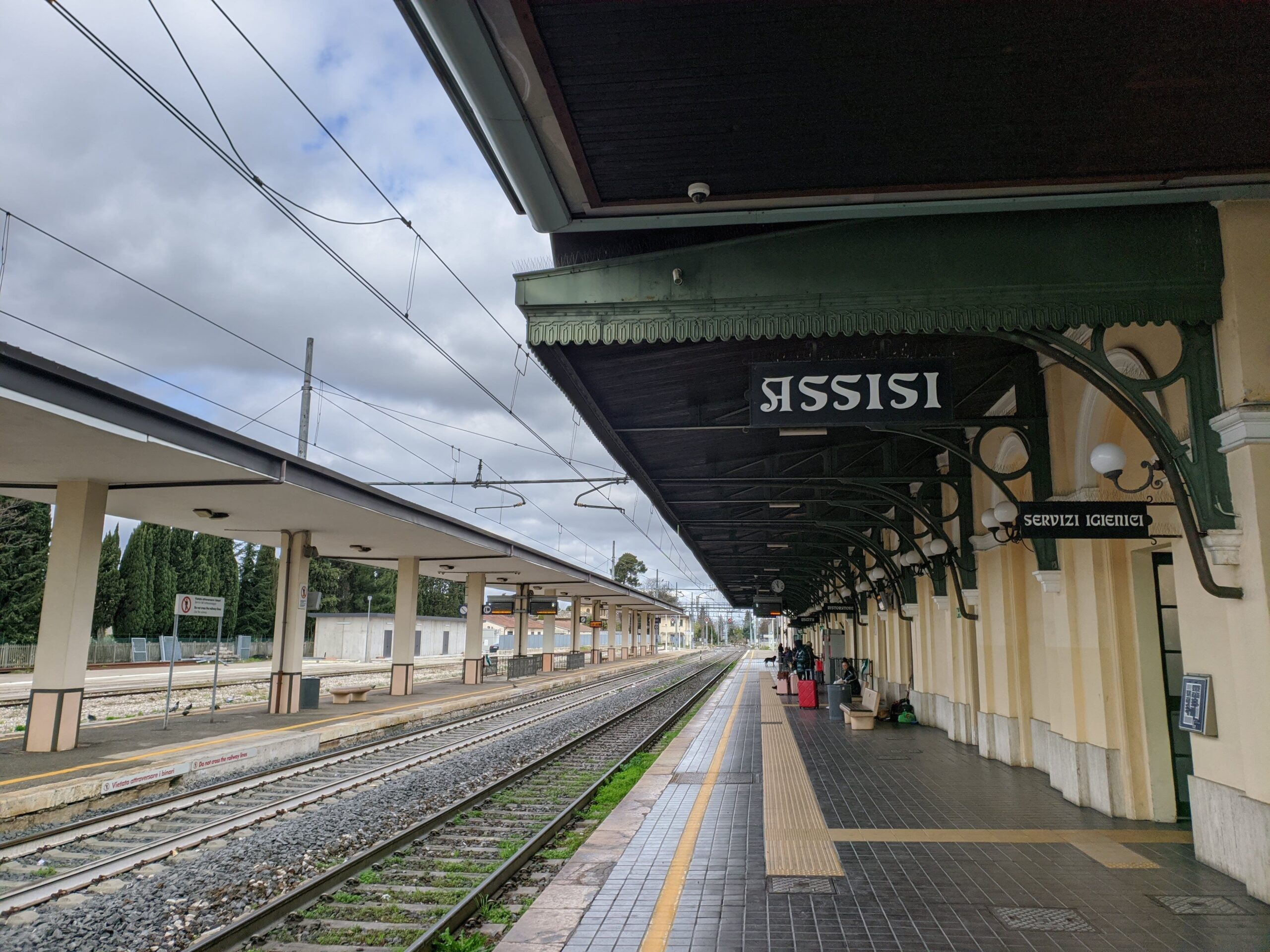 Plataforma da estação de trem de Assisi, com bancos vazios e um trem parado à distância, sob um céu nublado, indicando a chegada ou partida de Assis.