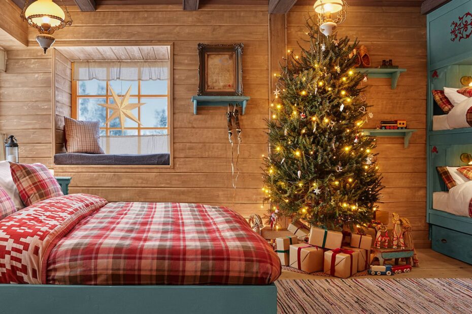 Quarto da Cabana do Papai Noel com cama de casal, árvore de Natal iluminada e beliches ao fundo em um ambiente rústico e festivo.