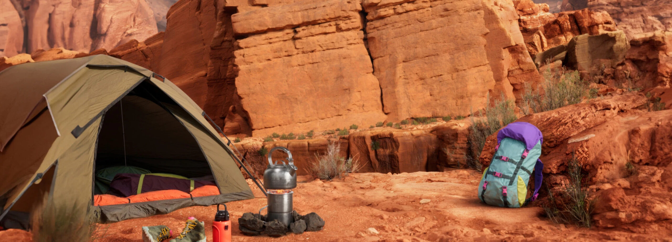 Uma tenda de camping marrom montada em um terreno arenoso com uma mochila colorida e um fogareiro com uma chaleira. O fundo mostra imponentes formações rochosas de cor vermelha sob um céu azul claro, evocando o espírito de aventura no deserto.