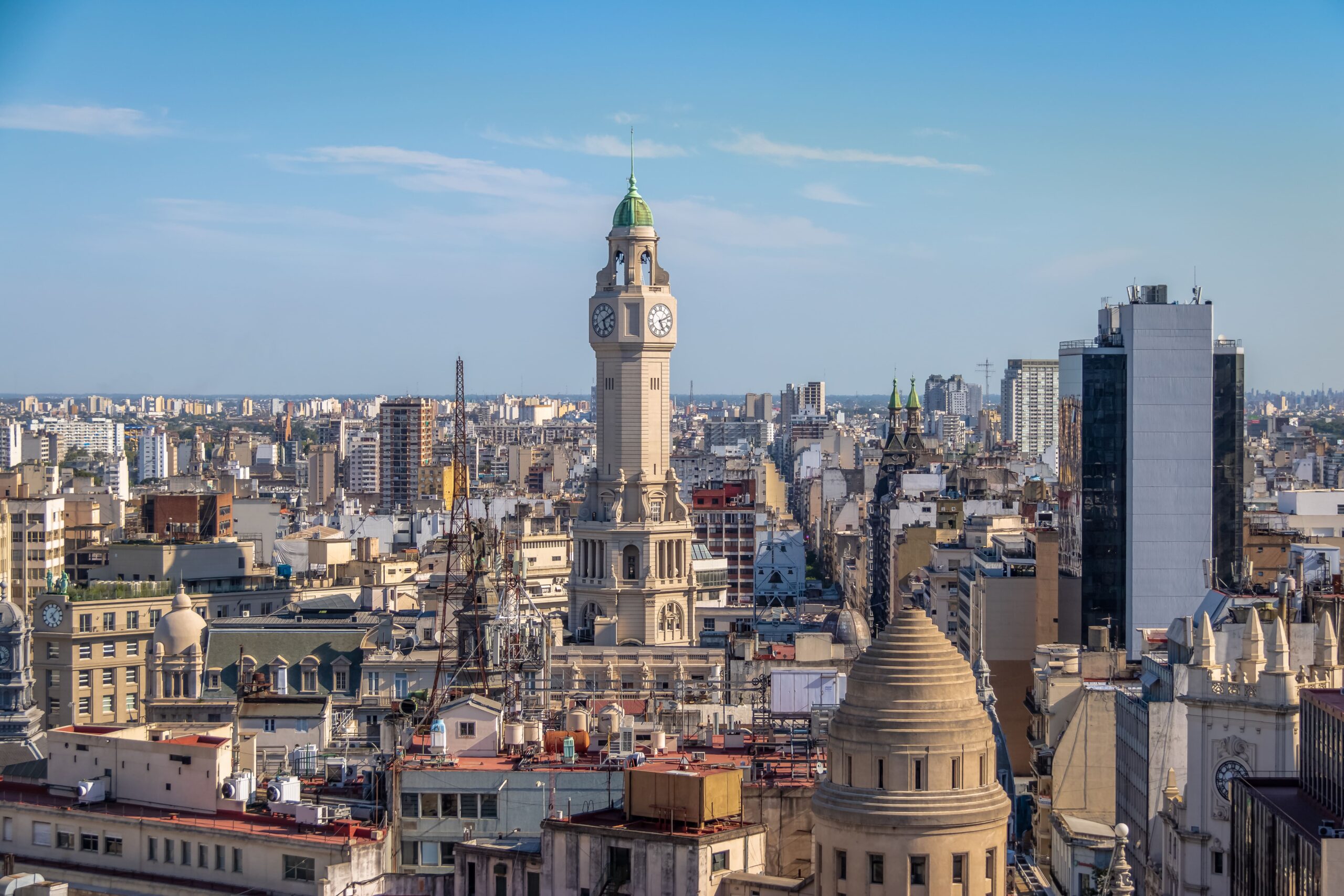 Vista aérea da cidade de Buenos Aires, mostrando a Torre da Legislatura e uma paisagem urbana variada, com edifícios de diferentes estilos arquitetônicos e alturas. O céu é claro, indicando um dia ensolarado.