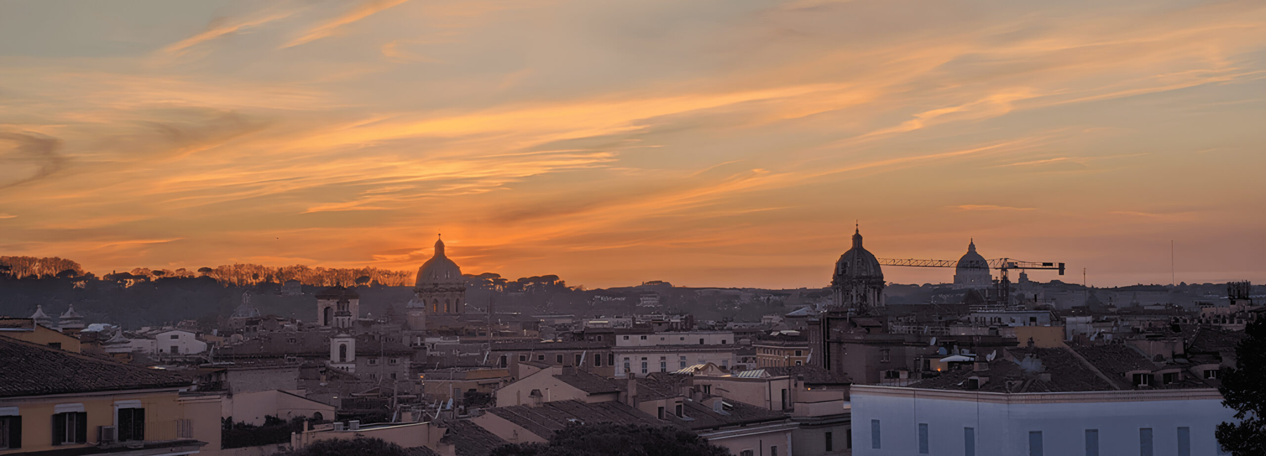 A imagem destaca a silhueta da cidade de Roma contra um céu vibrante de pôr do sol, com nuvens iluminadas por tons de laranja e amarelo. Os contornos das cúpulas e edifícios históricos são visíveis, oferecendo uma visão encantadora do horizonte da cidade ao anoitecer.