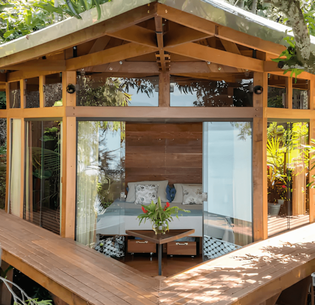 Uma cabana de madeira moderna e pequena situada em uma plataforma elevada entre a folhagem verde. A estrutura possui grandes janelas de vidro e uma decoração minimalista, integrando-se perfeitamente com o ambiente natural ao redor.