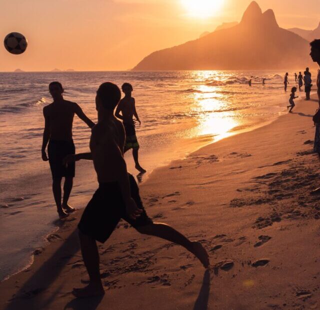 Silhueta de pessoas jogando futebol na praia ao pôr do sol. A cena acontece na praia do Rio de Janeiro, com o Morro Dois Irmãos ao fundo. As sombras das pessoas são projetadas na areia, onde se vê várias pegadas.