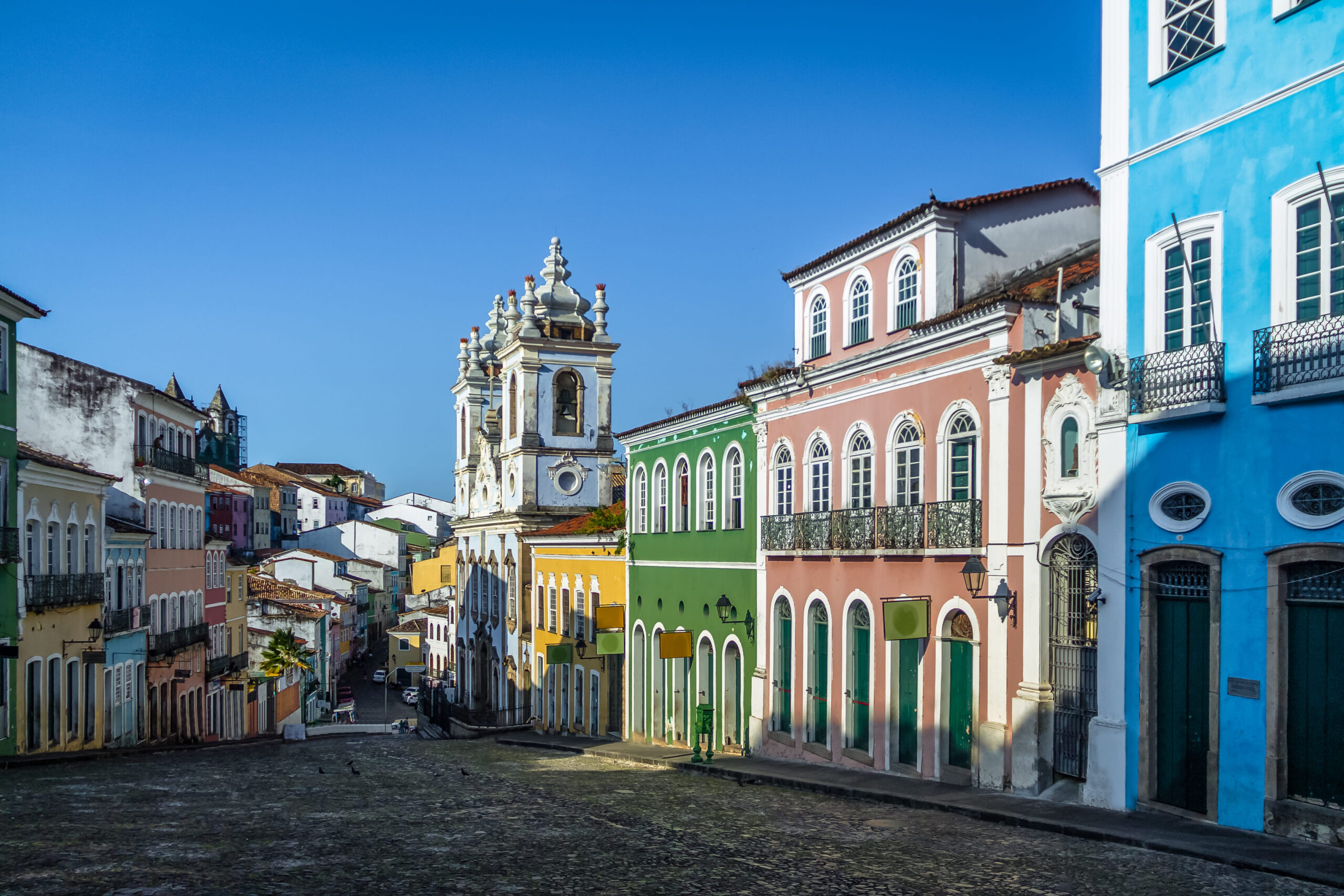 Colorida rua do Pelourinho em Salvador, Bahia, com casas históricas pintadas de diferentes cores. Uma igreja barroca com duas torres pode ser vista ao fundo. O céu é azul e a rua está vazia, criando uma atmosfera tranquila.
