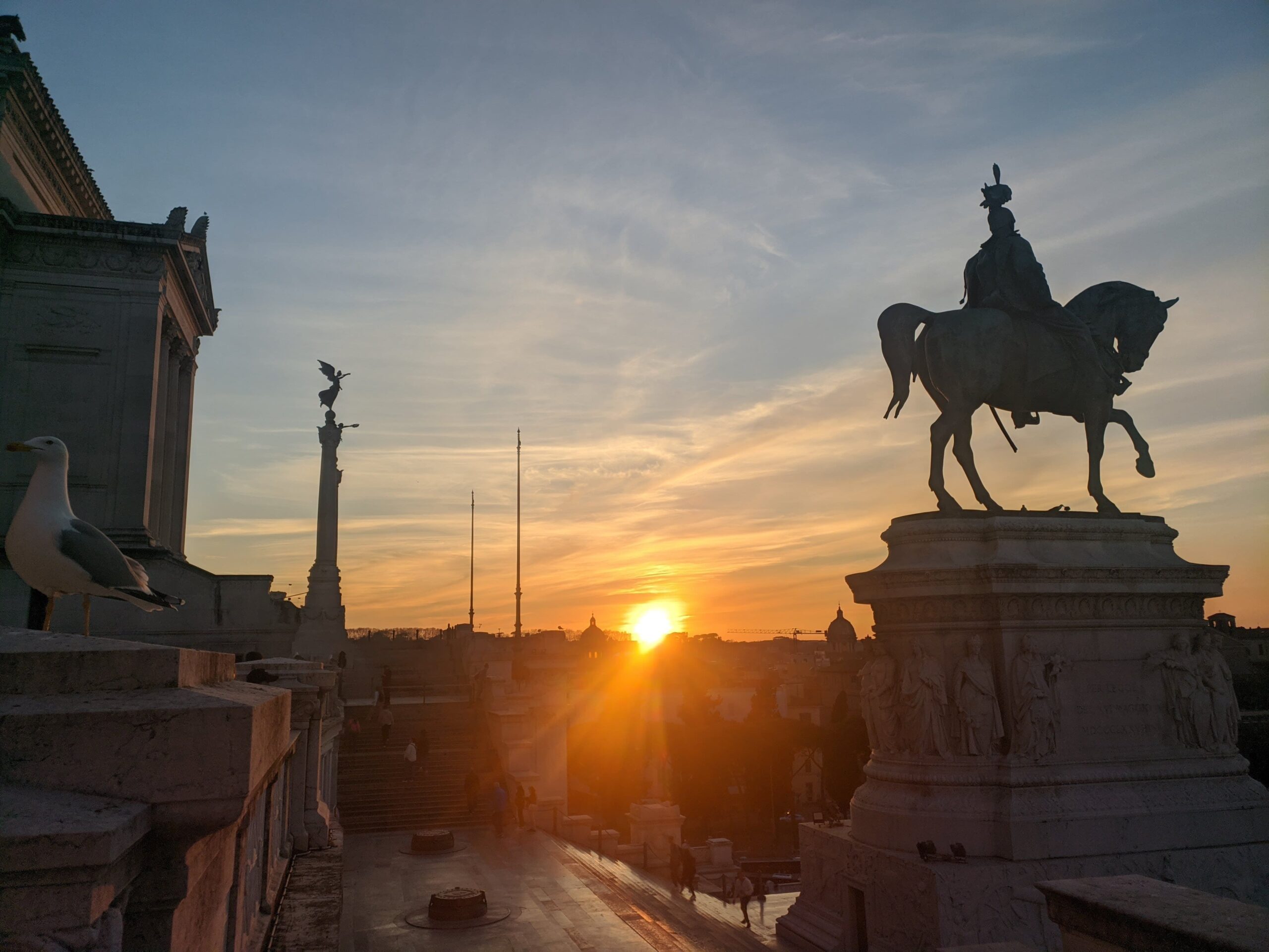 A imagem apresenta uma composição artística de Roma com uma estátua equestre em primeiro plano, um pássaro pousado à esquerda e o sol poente ao fundo, criando linhas de luz e sombra que realçam a atmosfera histórica e a beleza natural da cidade.