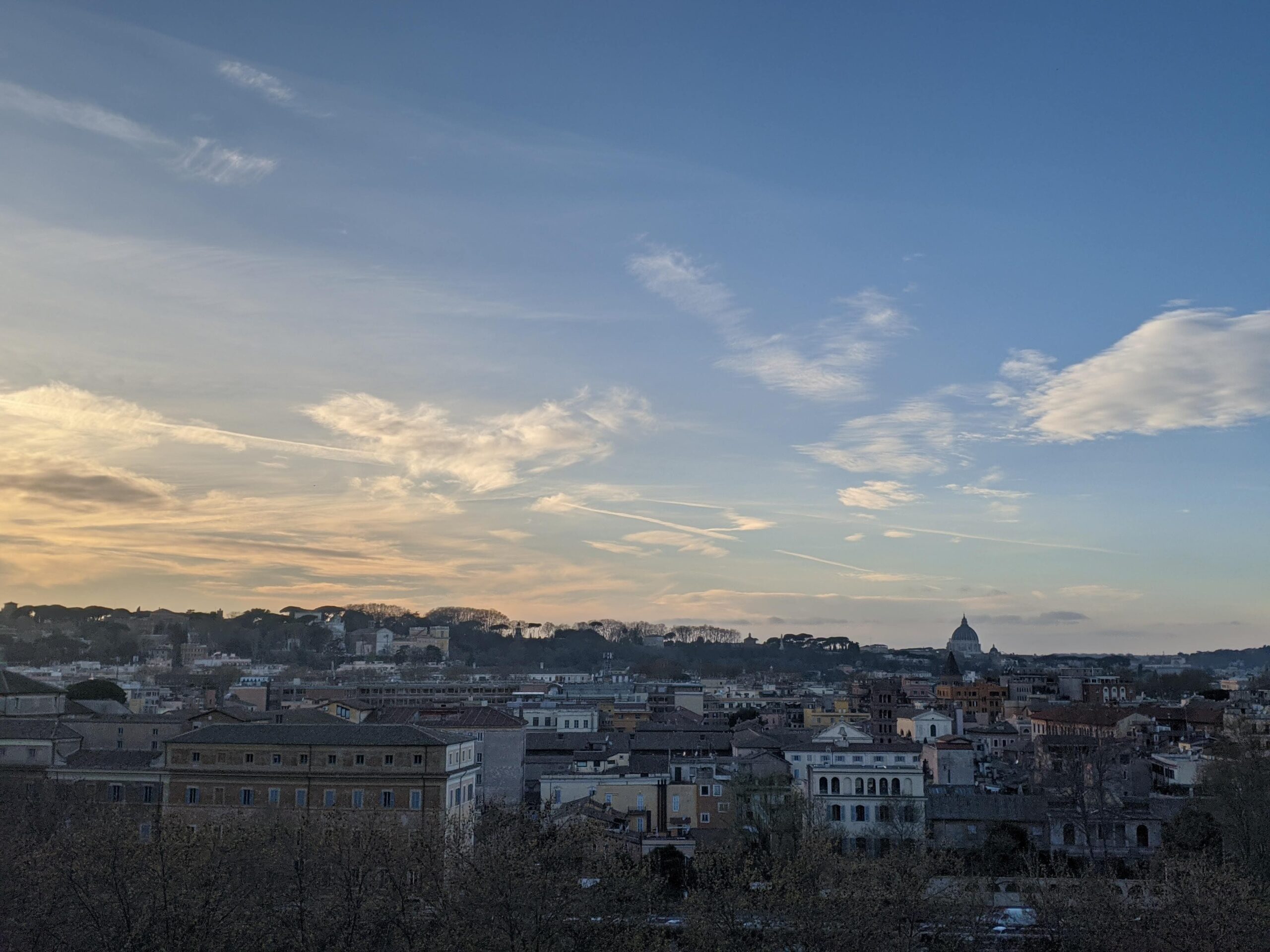 A imagem apresenta uma vista panorâmica da cidade de Roma ao entardecer, com inúmeras luzes já acesas, iluminando o tecido urbano. O céu acima da linha do horizonte mostra gradientes de azul e laranja, enquanto árvores despidas de folhas em primeiro plano adicionam um contraste dramático.