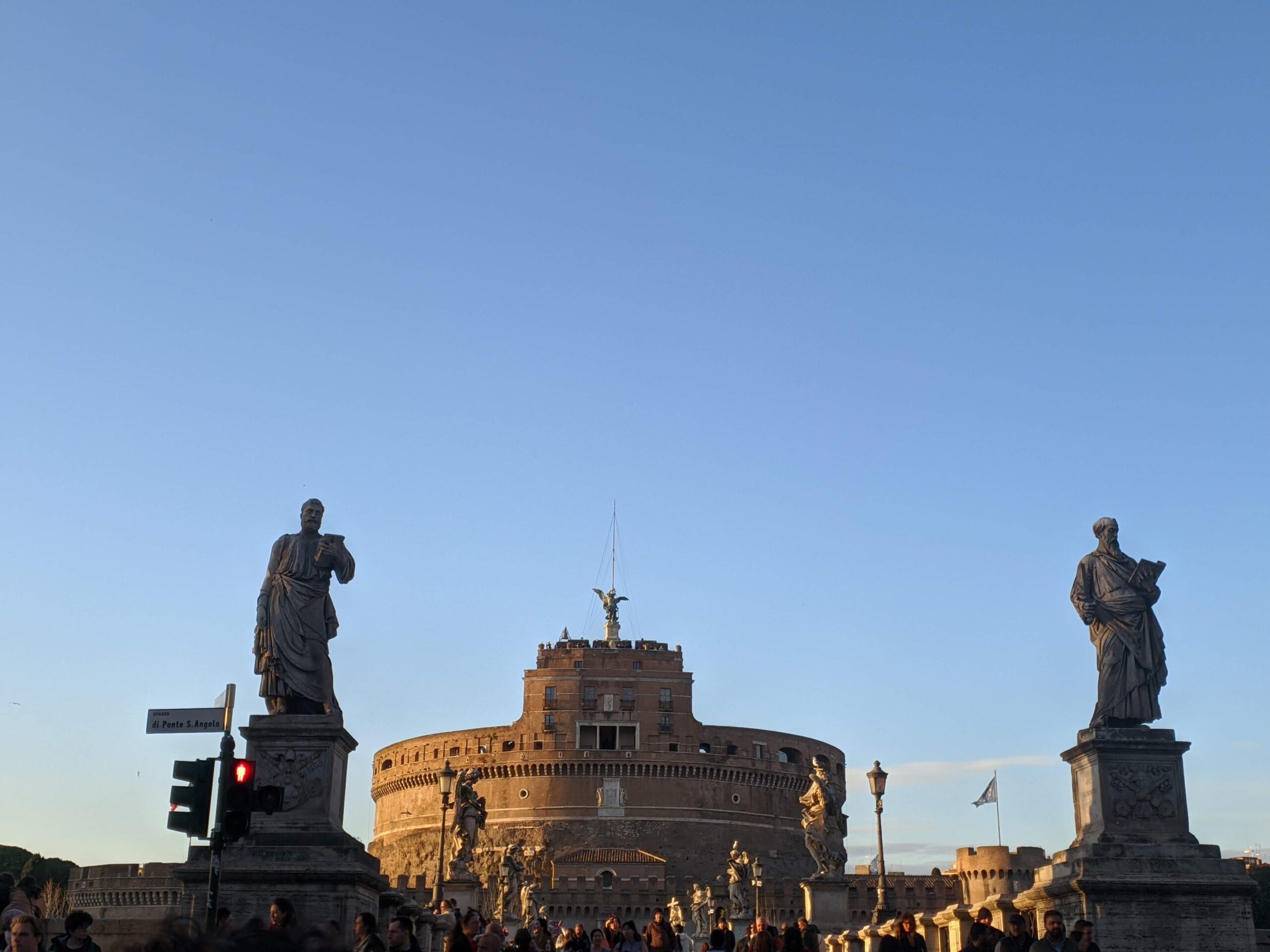 A imagem exibe uma vista ampla da cidade de Roma ao pôr do sol, com edifícios residenciais e monumentos espalhados por colinas suaves. O céu está pintado com nuvens finas e a luz dourada do sol poente, destacando as silhuetas dos pinheiros e das cúpulas.