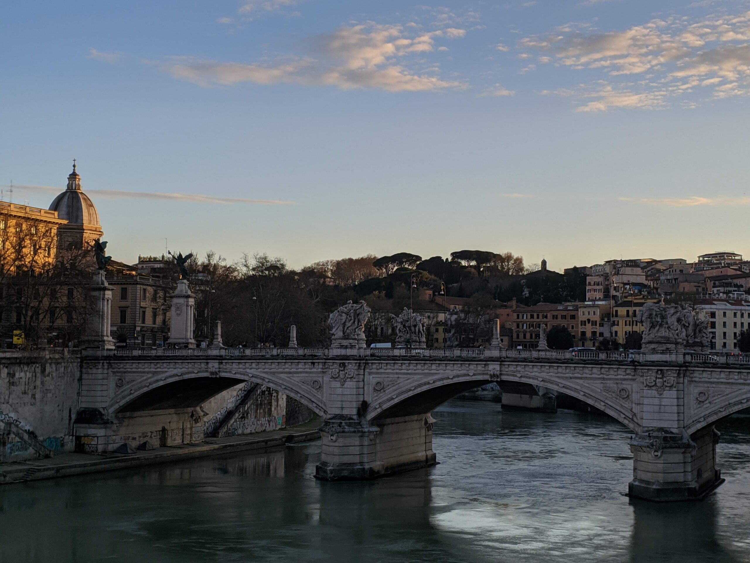 A imagem mostra uma ponte de pedra ornamentada com estátuas e balaustradas atravessando um rio tranquilo, com edifícios históricos ao fundo sob um céu azul claro. As águas calmas e as sombras suaves indicam que o sol está se pondo em Roma.