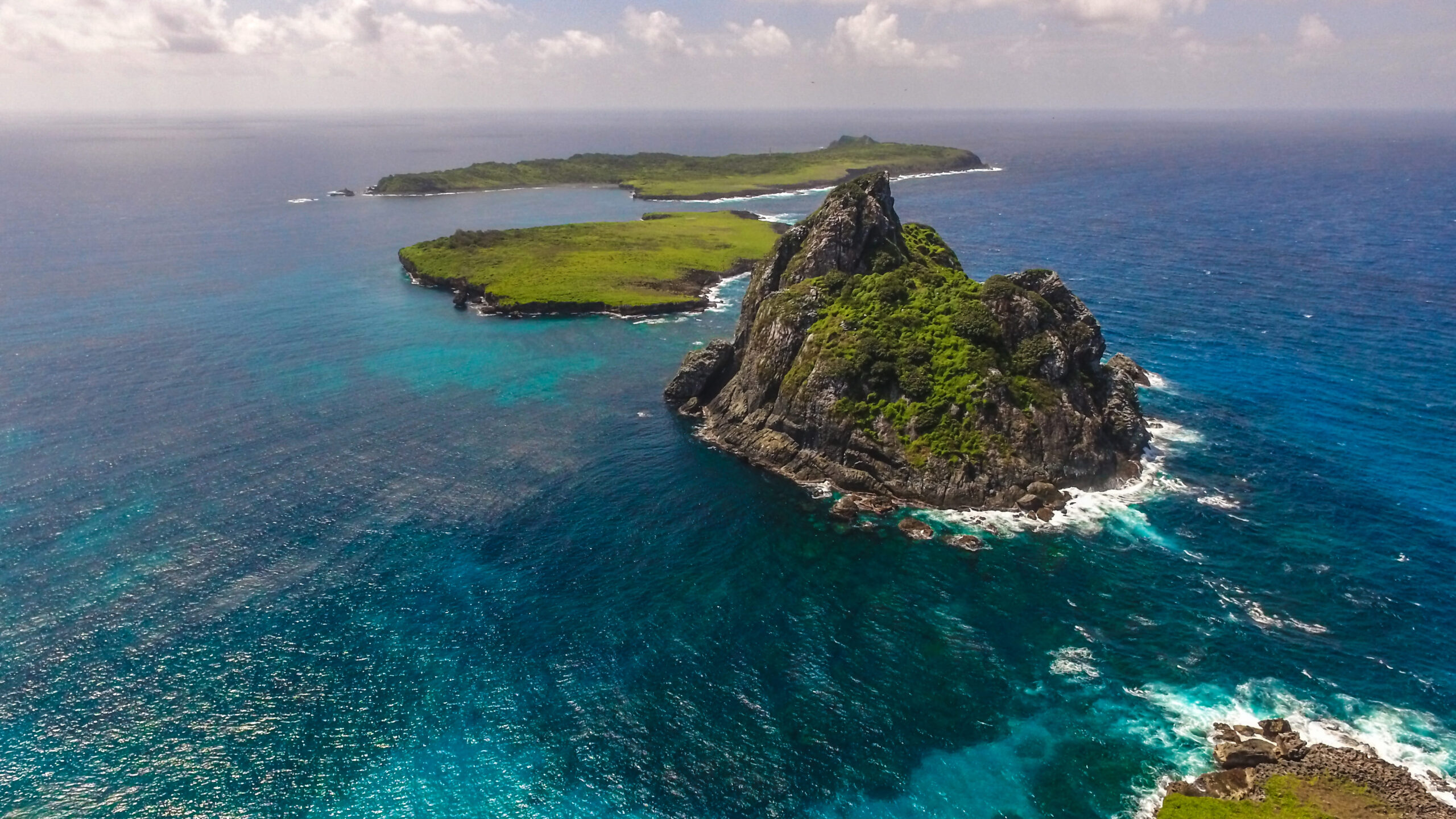 Vista aérea do Morro Dois Irmãos e da vegetação exuberante em Fernando de Noronha, destacando o contraste entre as águas azul-turquesa e as formações rochosas cobertas de verde, parte dos Patrimônios da UNESCO.