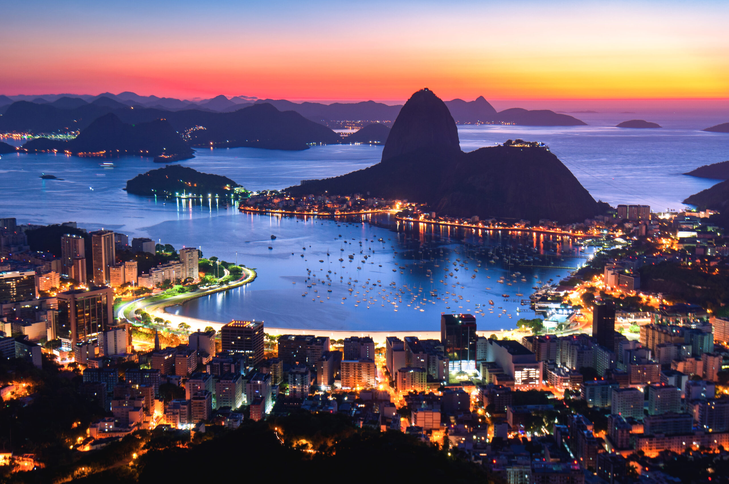 Vista noturna panorâmica do Rio de Janeiro com a Baía de Guanabara, o Pão de Açúcar ao fundo e as luzes da cidade refletindo na água, exemplificando a beleza natural e urbana do Patrimônio Mundial da UNESCO.