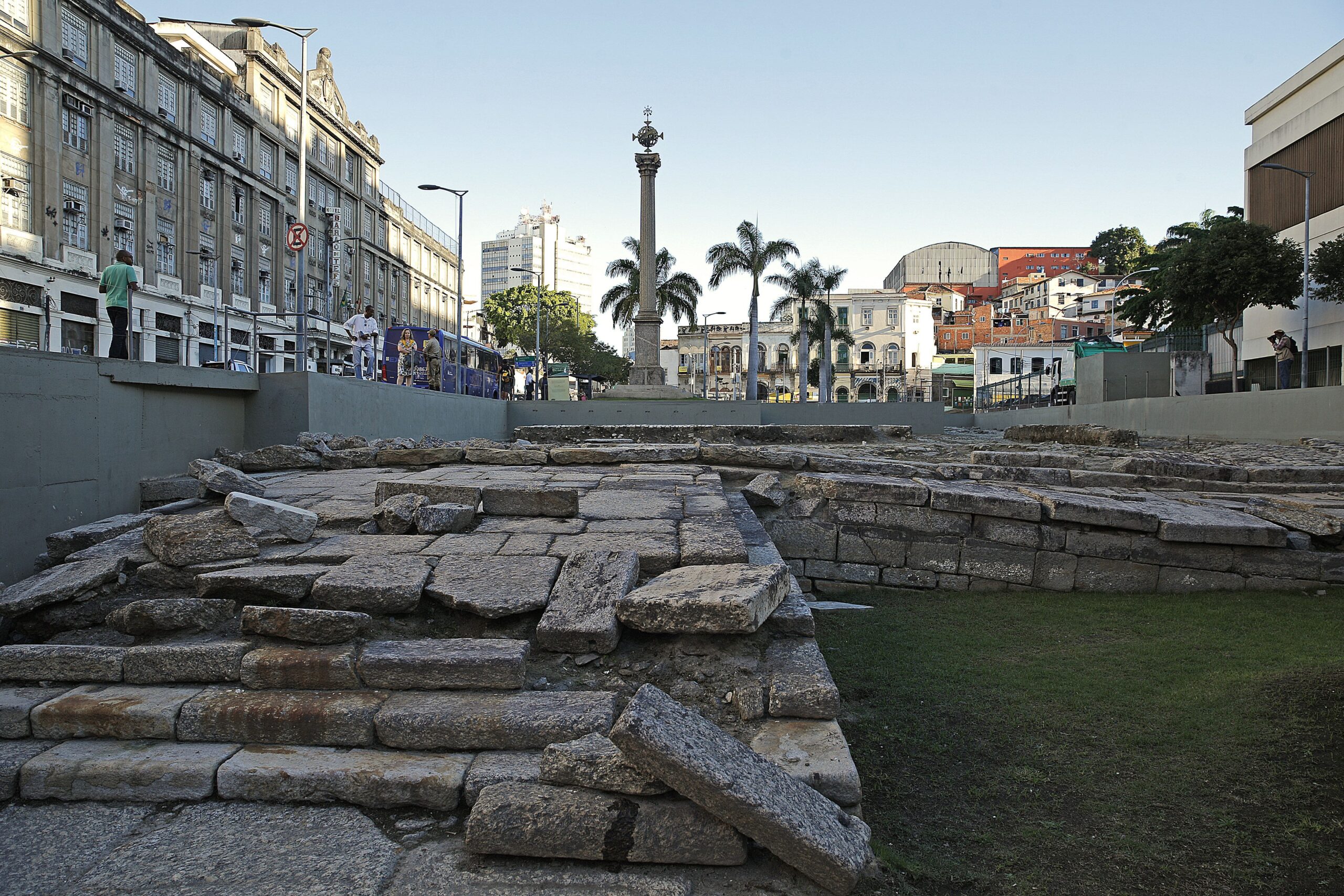 Ruínas históricas do Cais do Valongo no Rio de Janeiro, Brasil, um local de memória da diáspora africana e do comércio de escravizados, com prédios contemporâneos ao fundo e pessoas caminhando nas proximidades, refletindo a integração entre passado e presente em um Patrimônio Mundial da UNESCO.