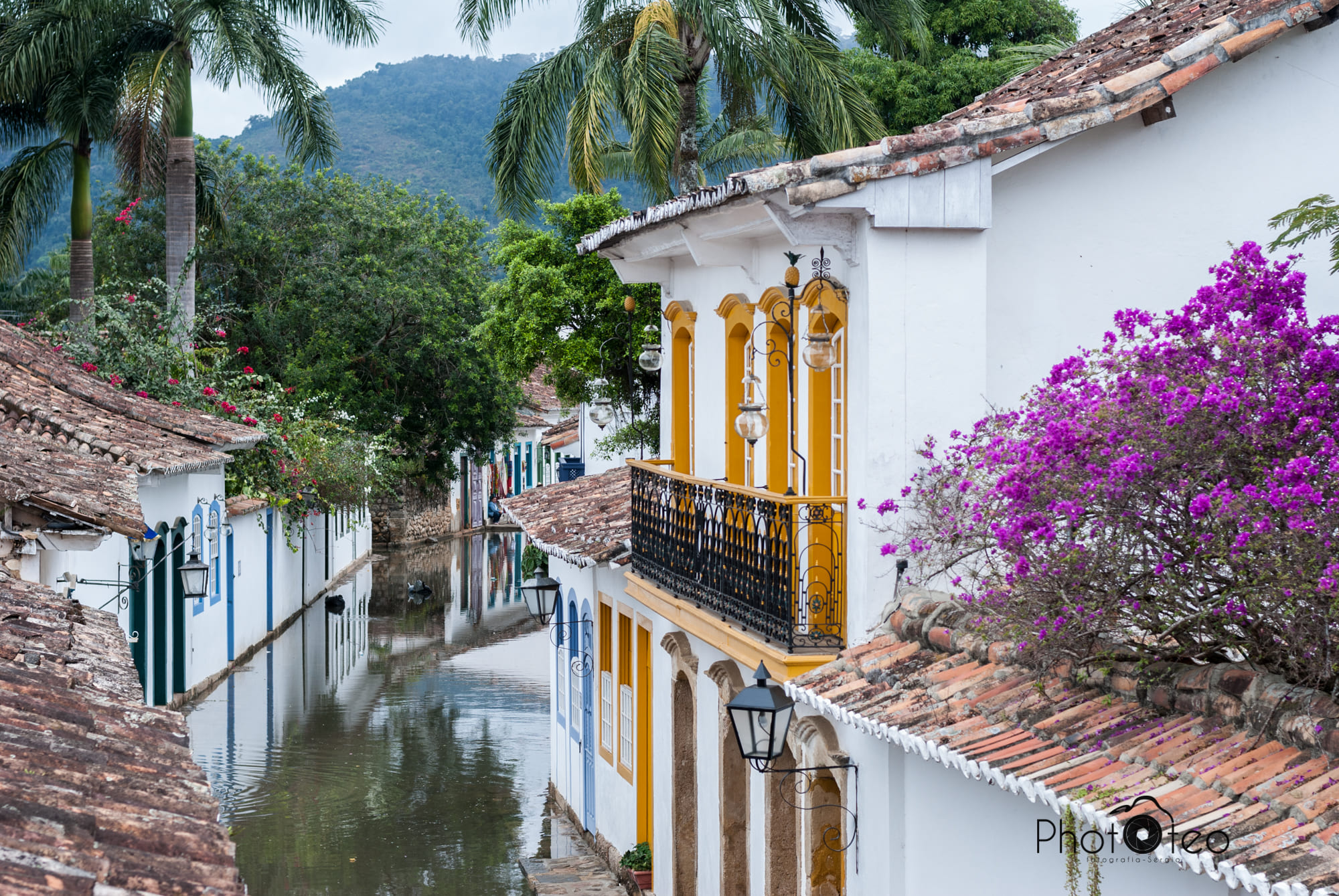 Rua histórica de Paraty alagada, com águas calmas refletindo as fachadas brancas dos edifícios coloniais e uma floração vibrante de buganvílias roxas, exemplificando a beleza e o charme deste Patrimônio Mundial da UNESCO.