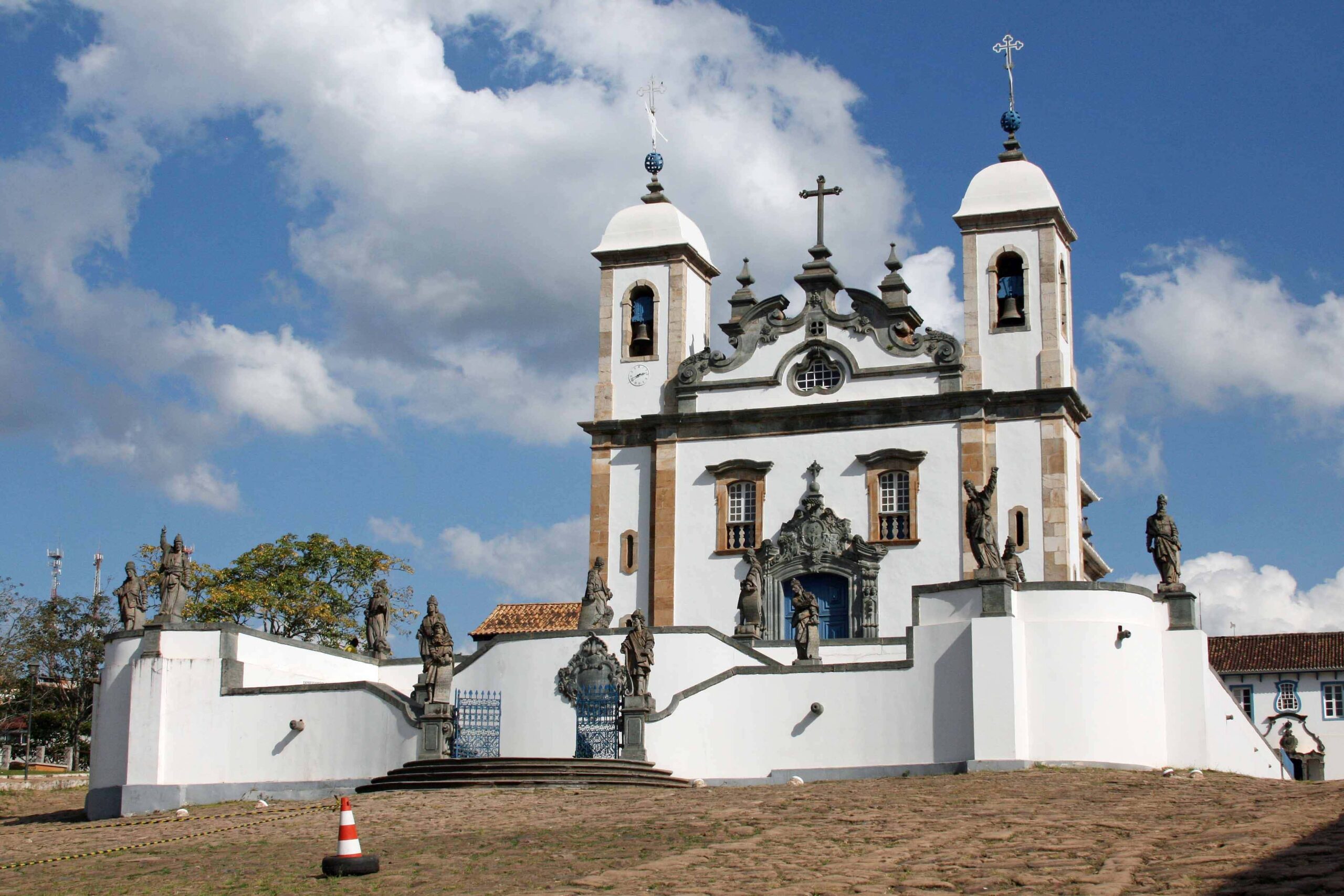 Igreja de São Francisco de Assis em Congonhas, mostrando as esculturas dos profetas de Aleijadinho em pedra-sabão, parte do Santuário do Bom Jesus de Matosinhos, sob um céu azul com nuvens.