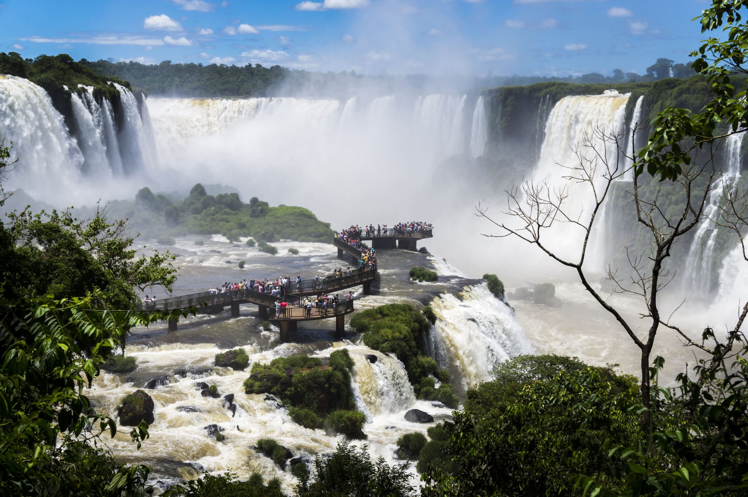 Vista das majestosas Cataratas do Iguaçu com vastas quedas d'água cercadas por vegetação exuberante e um mirante cheio de visitantes admirando a paisagem.