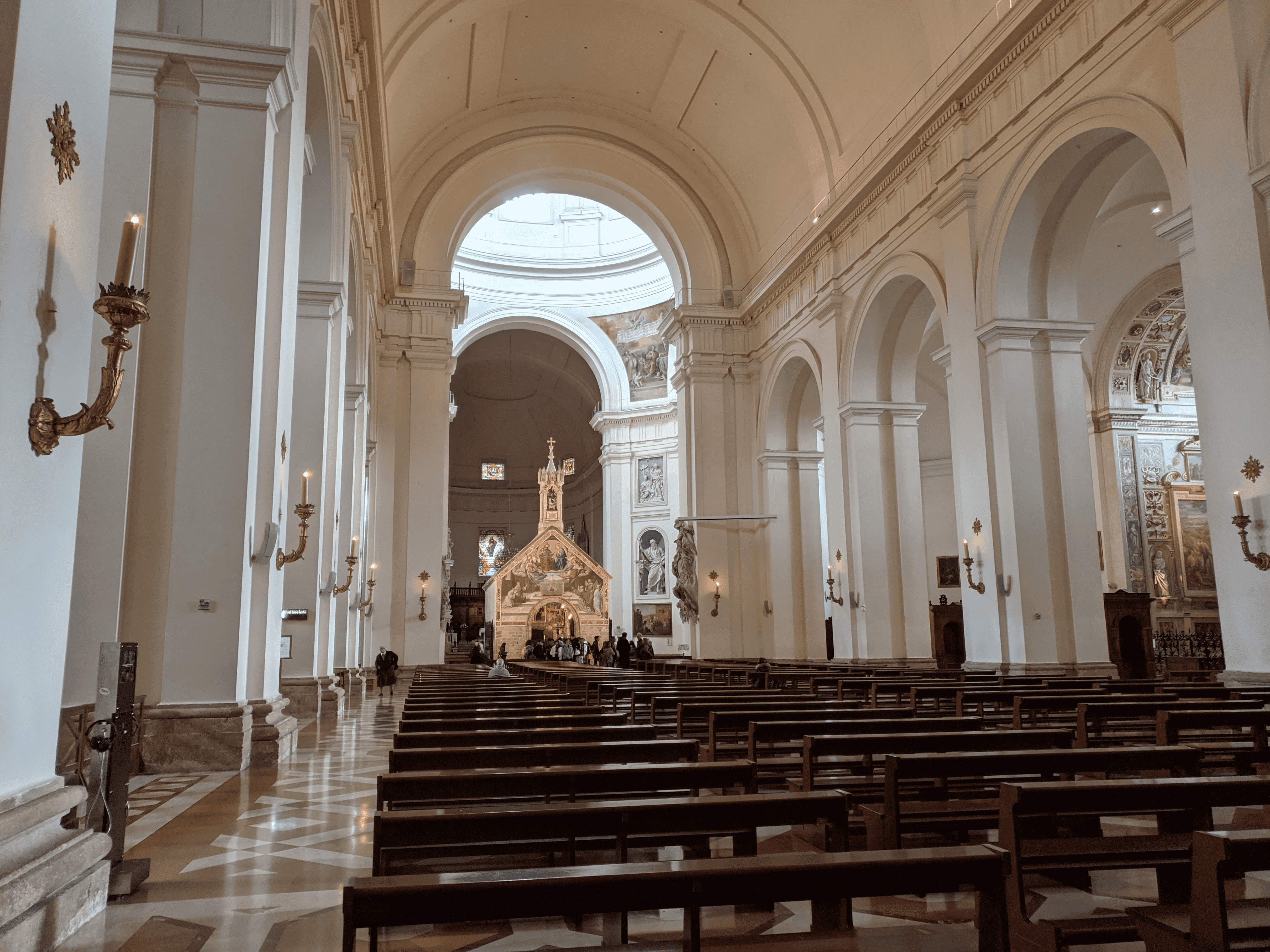 Interior de uma igreja com longos bancos de madeira alinhados, paredes e colunas brancas, e um altar ao fundo sob uma abóbada iluminada.