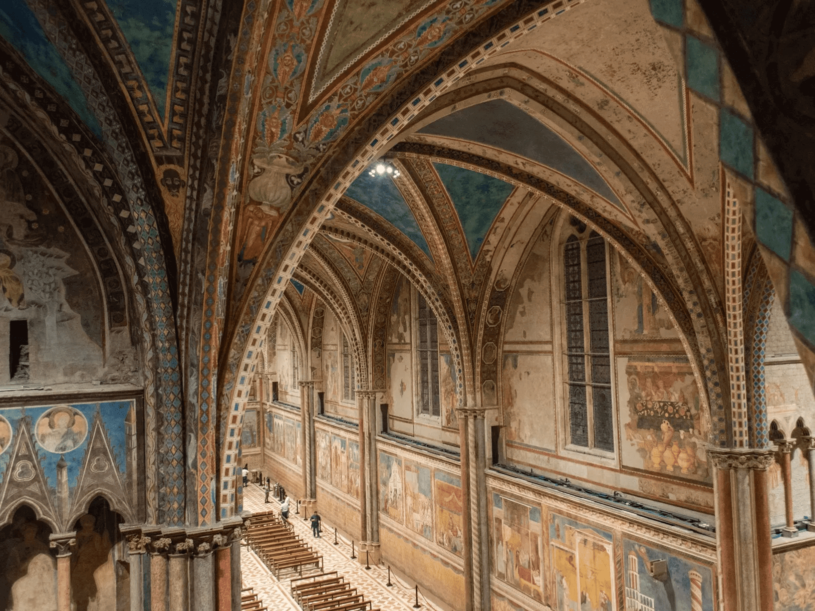 Interior da Basílica de São Francisco de Assis, em sua parte maior, ricamente decorada com afrescos nas paredes e tetos em arco, com detalhes em azul e dourado, e bancos de madeira dispostos no centro.