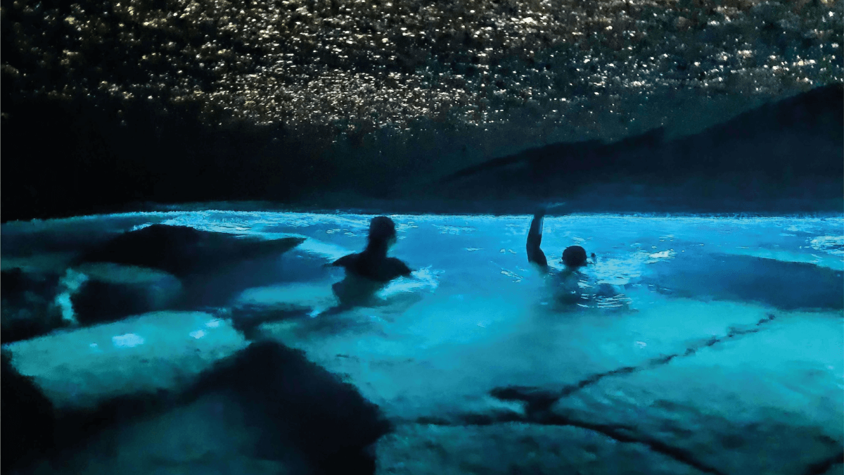 Cena de dentro da Gruta do Acaiá, um dos mais bonitos passeios na Ilha Grande, duas pessoas estão mergulhando em águas azul fluorescente, que é coberta por uma grande pedra.