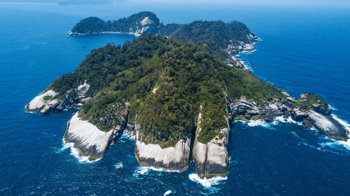 Vista aérea de uma ilha tropical com penhascos íngremes e vegetação exuberante. O oceano azul profundo circunda a ilha, com pequenas ondas batendo contra as rochas.