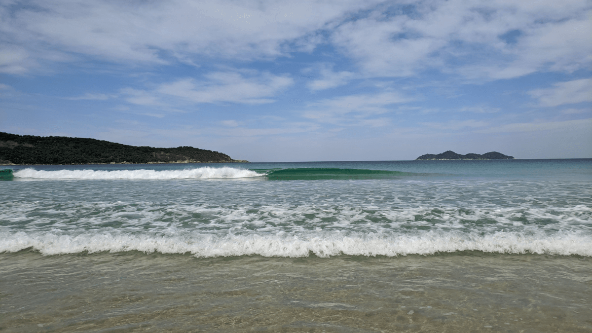 Praia tranquila com ondas suaves chegando à costa, cercada por vegetação verde e montanhas ao fundo sob um céu claro.