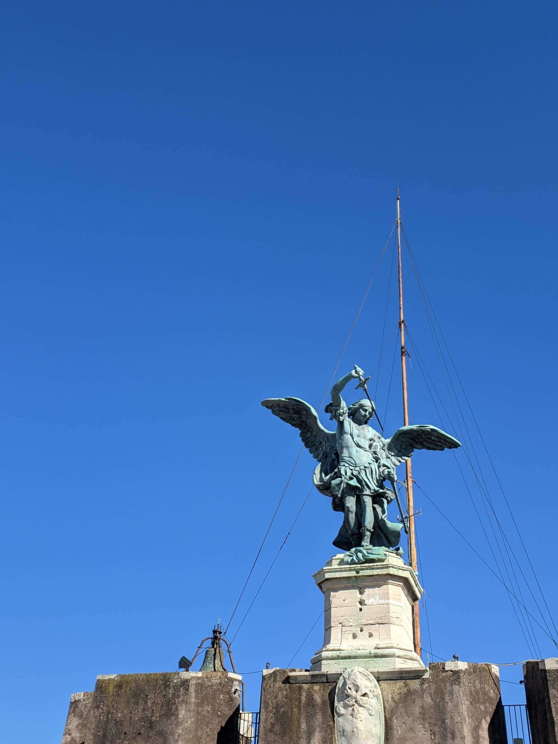 Estátua em bronze do Arcanjo Miguel com espada e asas, localizada no topo do Castelo de Santo Ângelo contra um céu azul claro.