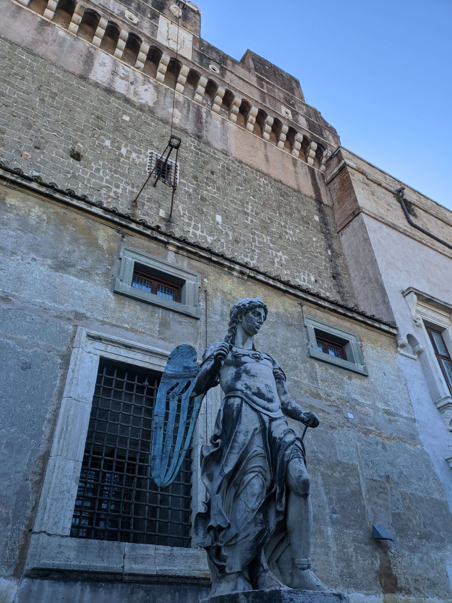 Estátua do Arcanjo Miguel com escudo e espada, no Castelo de Santo Ângelo, mostrando detalhes da arquitetura antiga ao fundo.