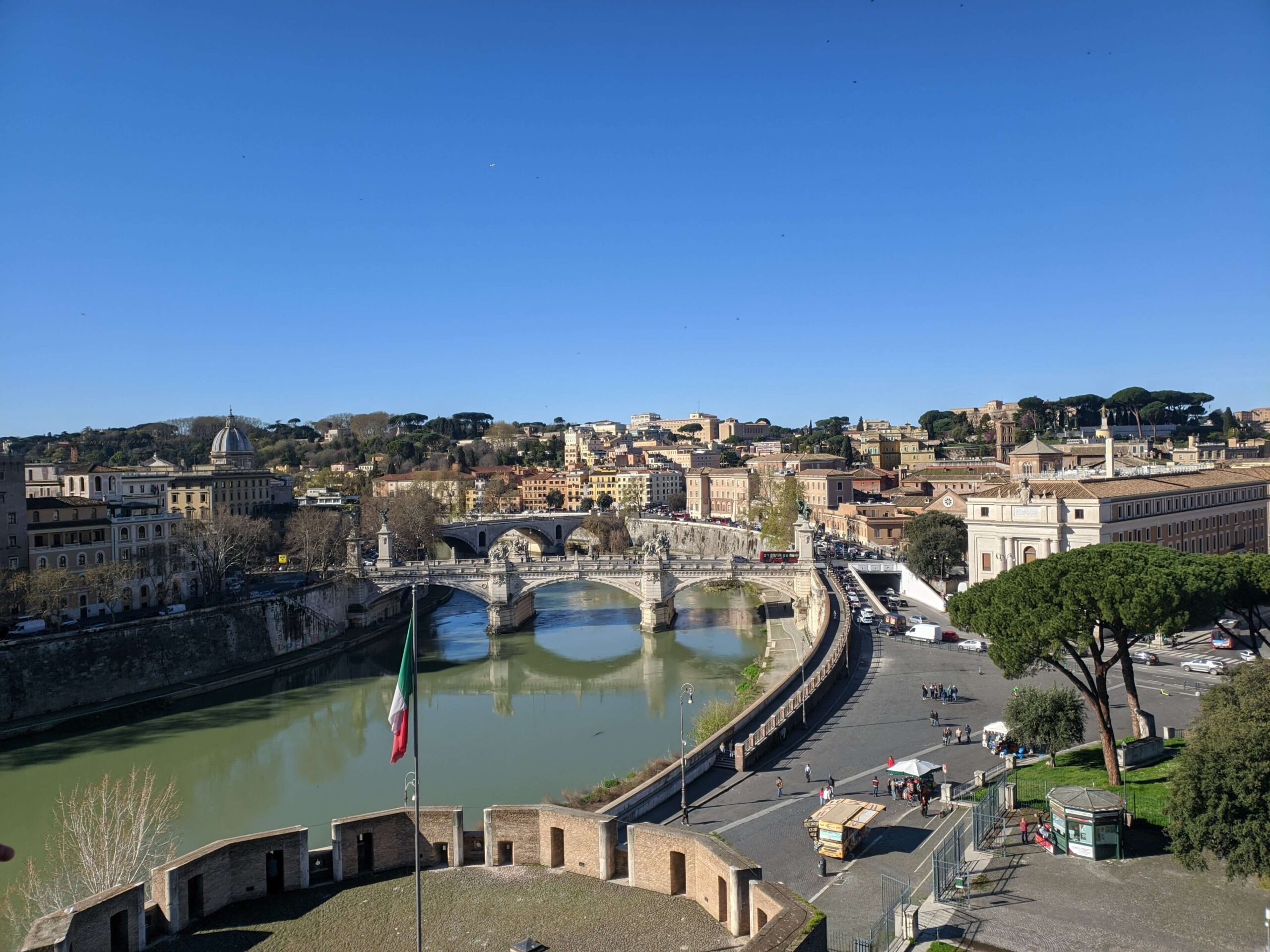 Vista panorâmica diurna do Rio Tibre e da Ponte Sant'Angelo com o centro de Roma ao fundo, sob um céu claro.