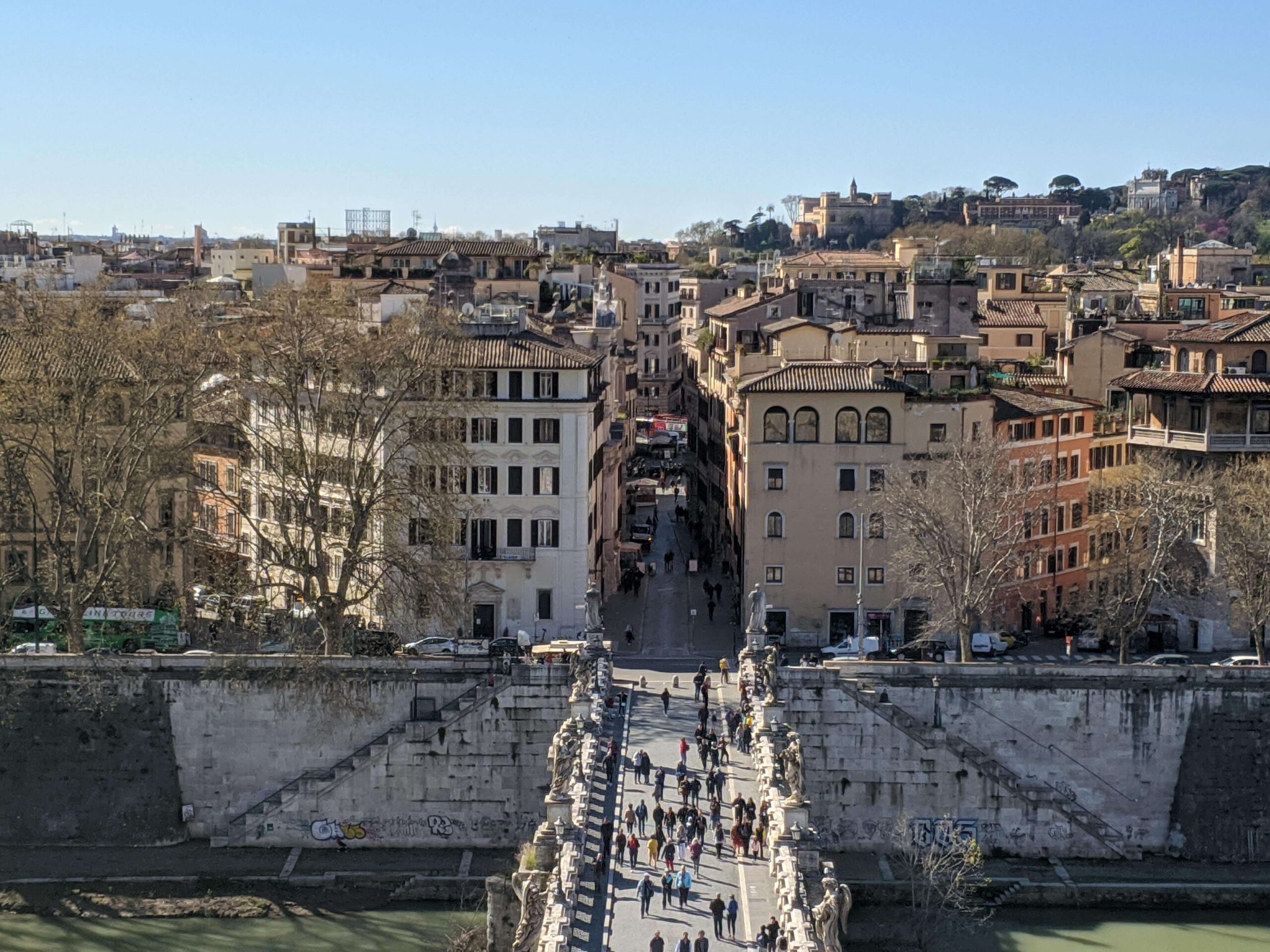Perspectiva mais próxima da Ponte Sant'Angelo cheia de estátuas, com a cidade de Roma ao fundo, sob uma iluminação clara.