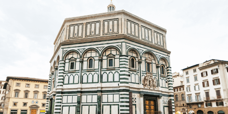 Imagem do Batistério de São João em Florença, com sua notável fachada de mármore branco e verde, mostrando detalhes das portas e esculturas que adornam sua arquitetura exterior.