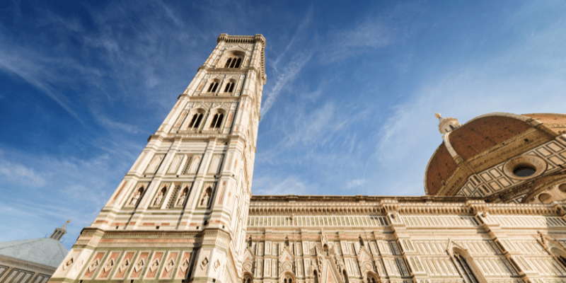 Imagem do Campanário do Duomo de Florença, parte do complexo da catedral, mostrando sua estrutura vertical e a rica decoração de mármore, sob um céu azul claro.