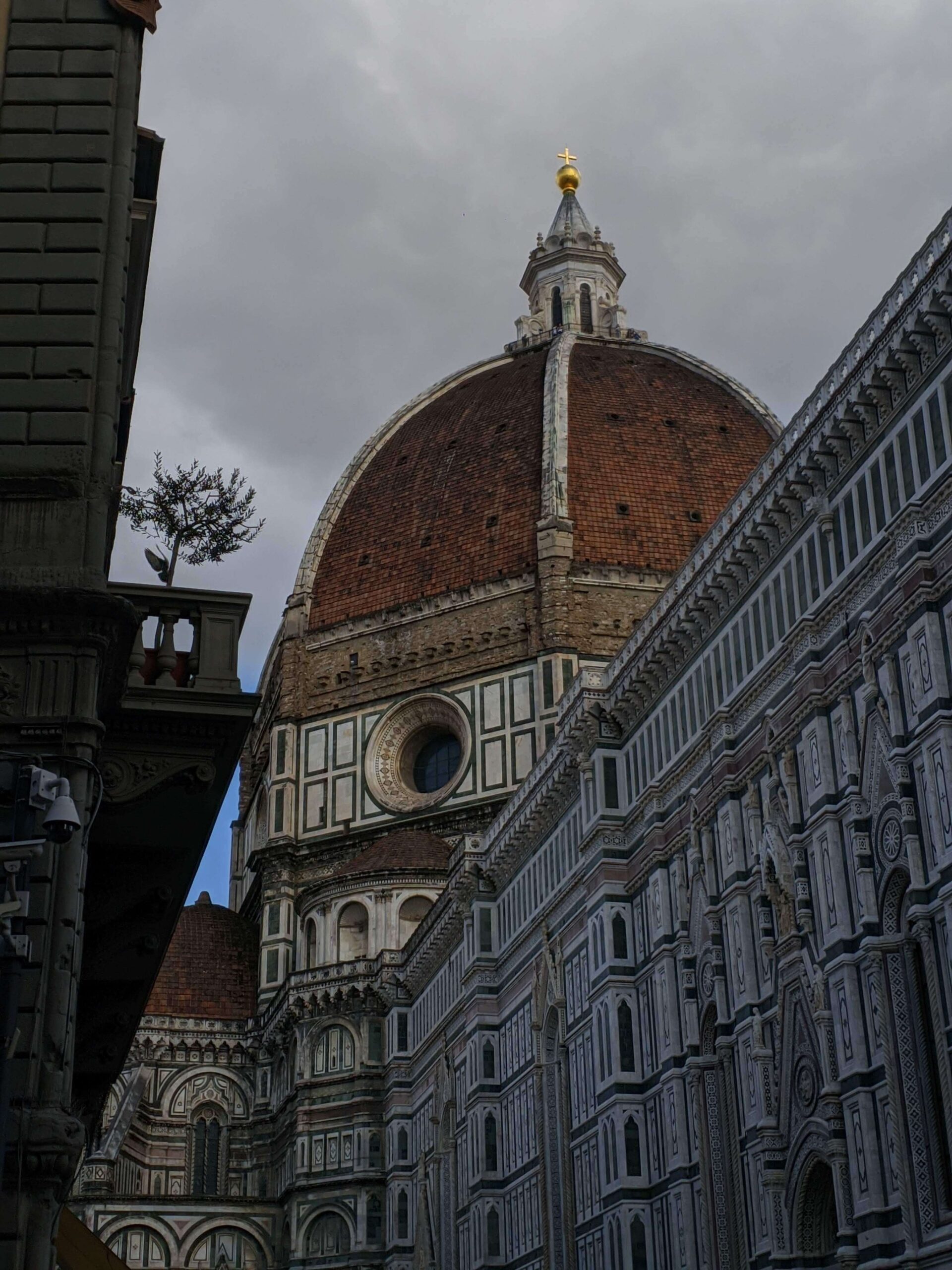 Visão da cúpula do Duomo de Florença entre edifícios históricos, sob um céu encoberto, destacando a harmonia entre a grandiosidade do Duomo e a escala urbana de Florença.