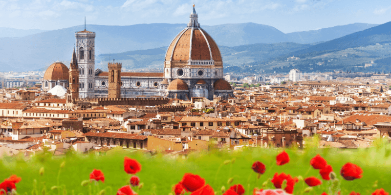Vista panorâmica do Duomo de Florença com a cidade ao fundo e flores em primeiro plano, destacando a cúpula e a torre do Campanário de Giotto.