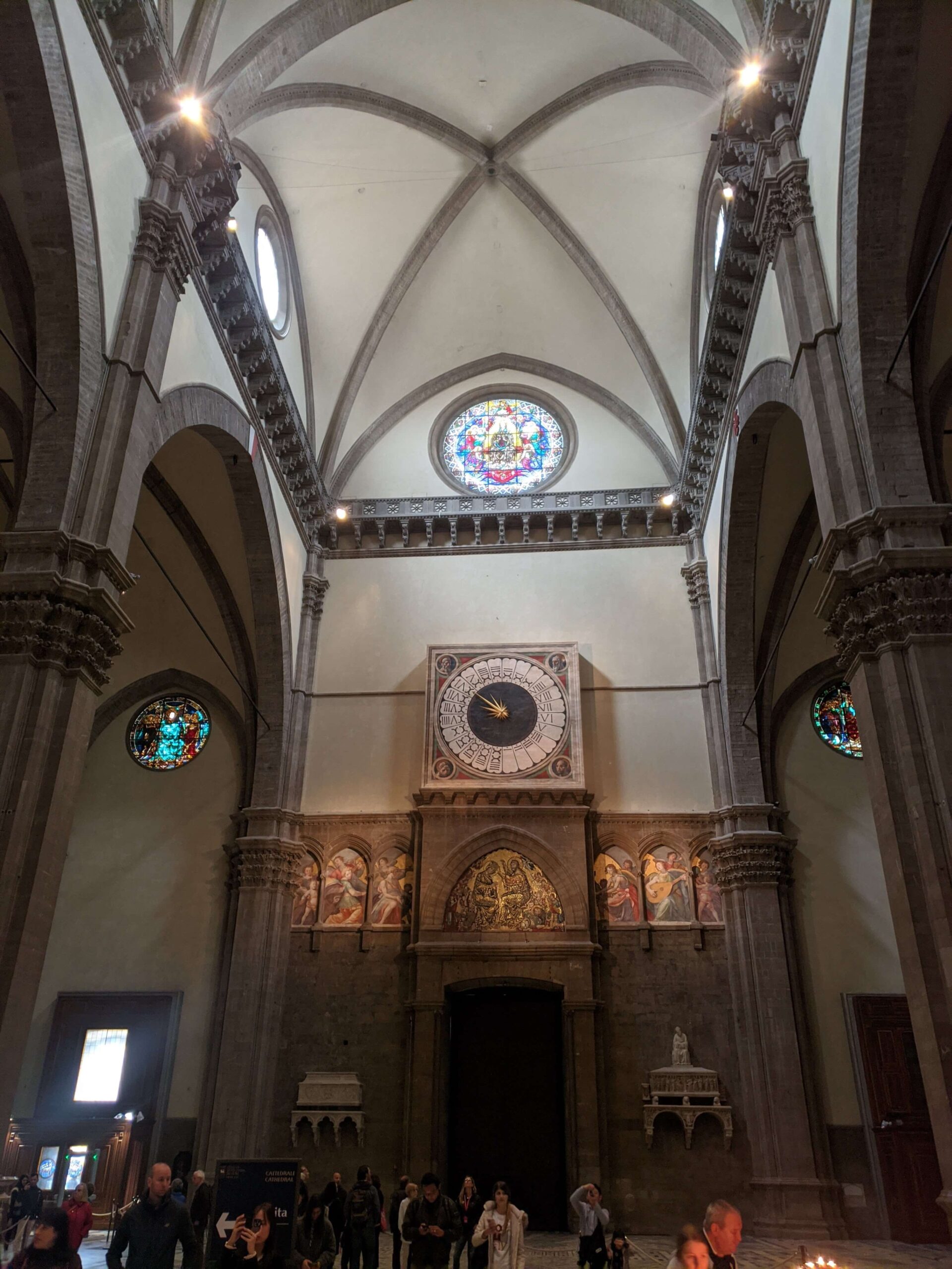 Interior do Duomo de Florença mostrando o vasto espaço da nave, com o relógio de parede acima da entrada principal e vitrais coloridos iluminando o ambiente.