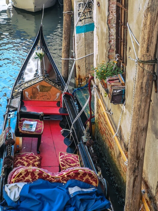 Uma gôndola tradicional amarrada a um pequeno cais em Veneza, com uma decoração interior vermelha e dourada e livros armazenados ao longo do exterior da embarcação.