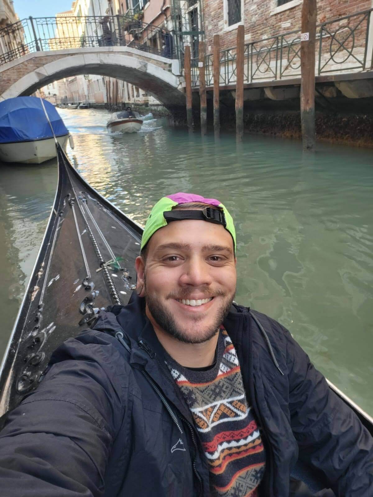 Um homem sorridente tira uma selfie enquanto passeia de gôndola em um canal de Veneza, com prédios históricos e uma ponte ao fundo.