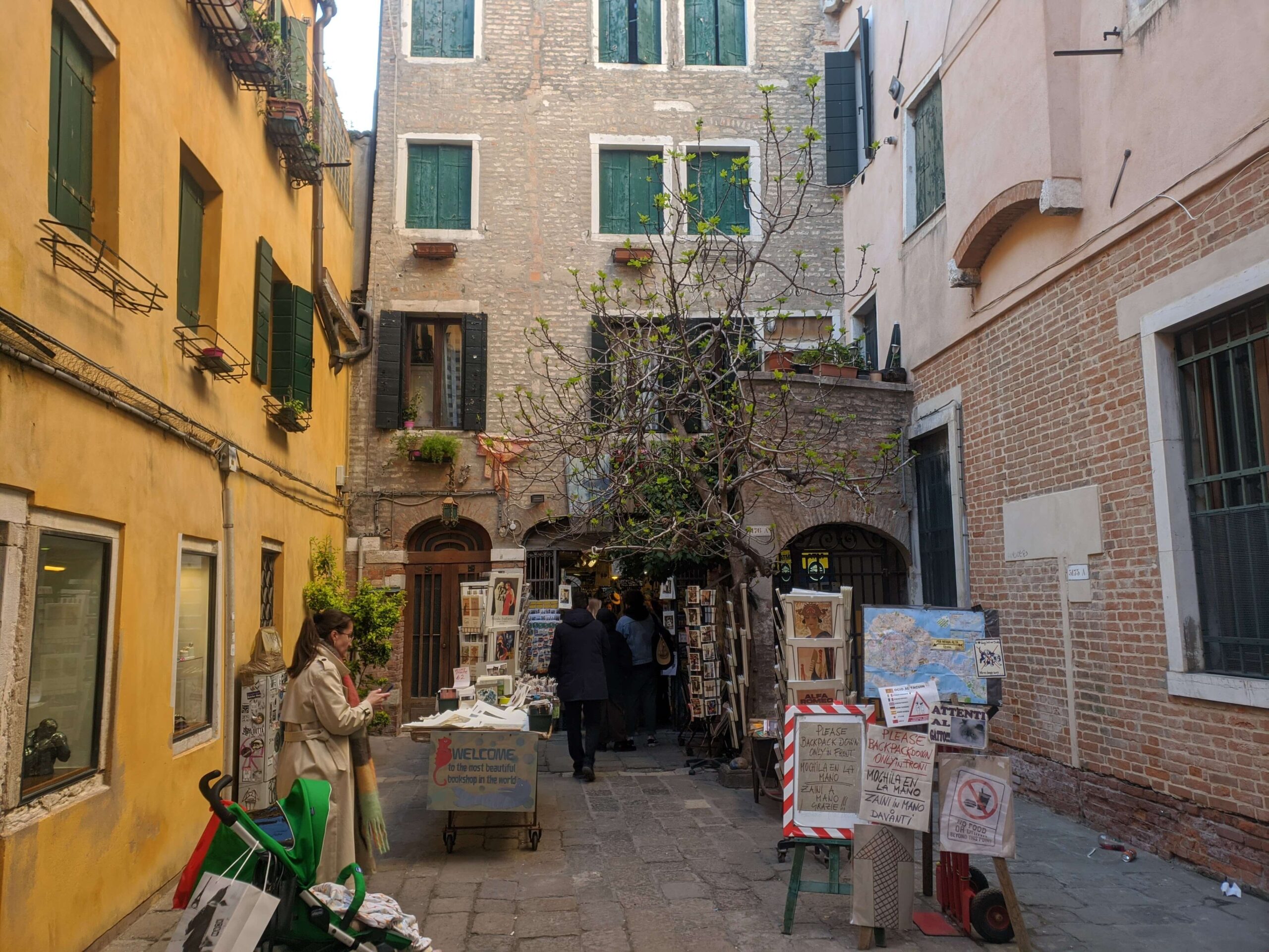 Uma rua de Veneza com prédios de tons quentes e uma pequena loja de arte ao ar livre. Pessoas caminham e exploram as mercadorias à venda, e há uma árvore que adiciona um toque de verde à cena urbana.