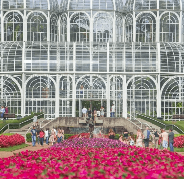 Uma imagem da icônica estufa do Jardim Botânico de Curitiba. A estrutura de metal e vidro é cercada por jardins vibrantes de flores pink. Turistas podem ser vistos passeando e apreciando as plantas ao redor da estufa, que é uma das principais atrações turísticas da cidade.