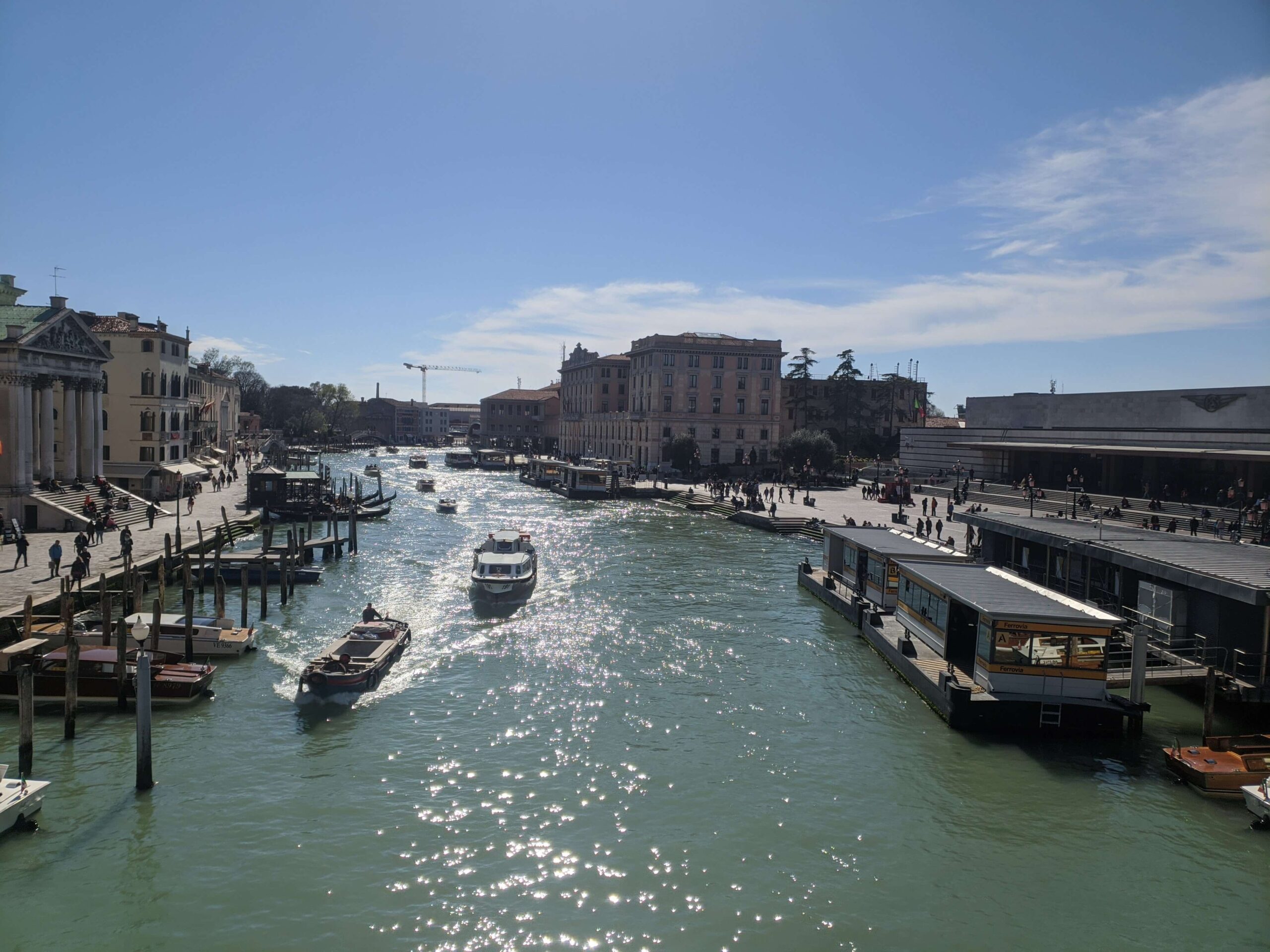 Uma visão ampla do Grande Canal de Veneza em um dia ensolarado, com embarcações transitando, pessoas caminhando ao longo das margens e arquitetura clássica veneziana em ambos os lados.