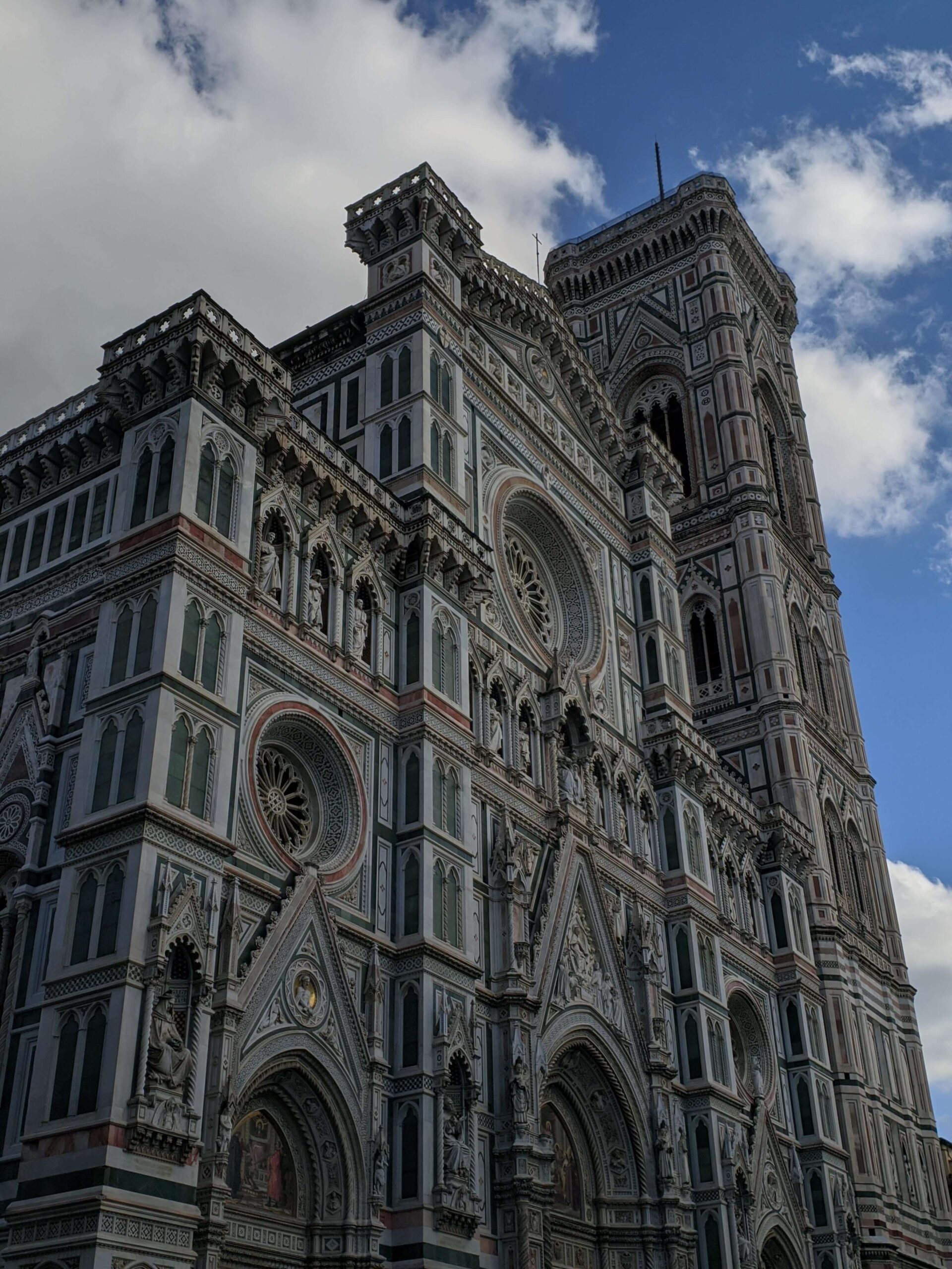 Fachada detalhada do Duomo de Florença sob um céu parcialmente nublado, destacando a intrincada arquitetura gótica, com seus mármores coloridos e a grande rosácea.